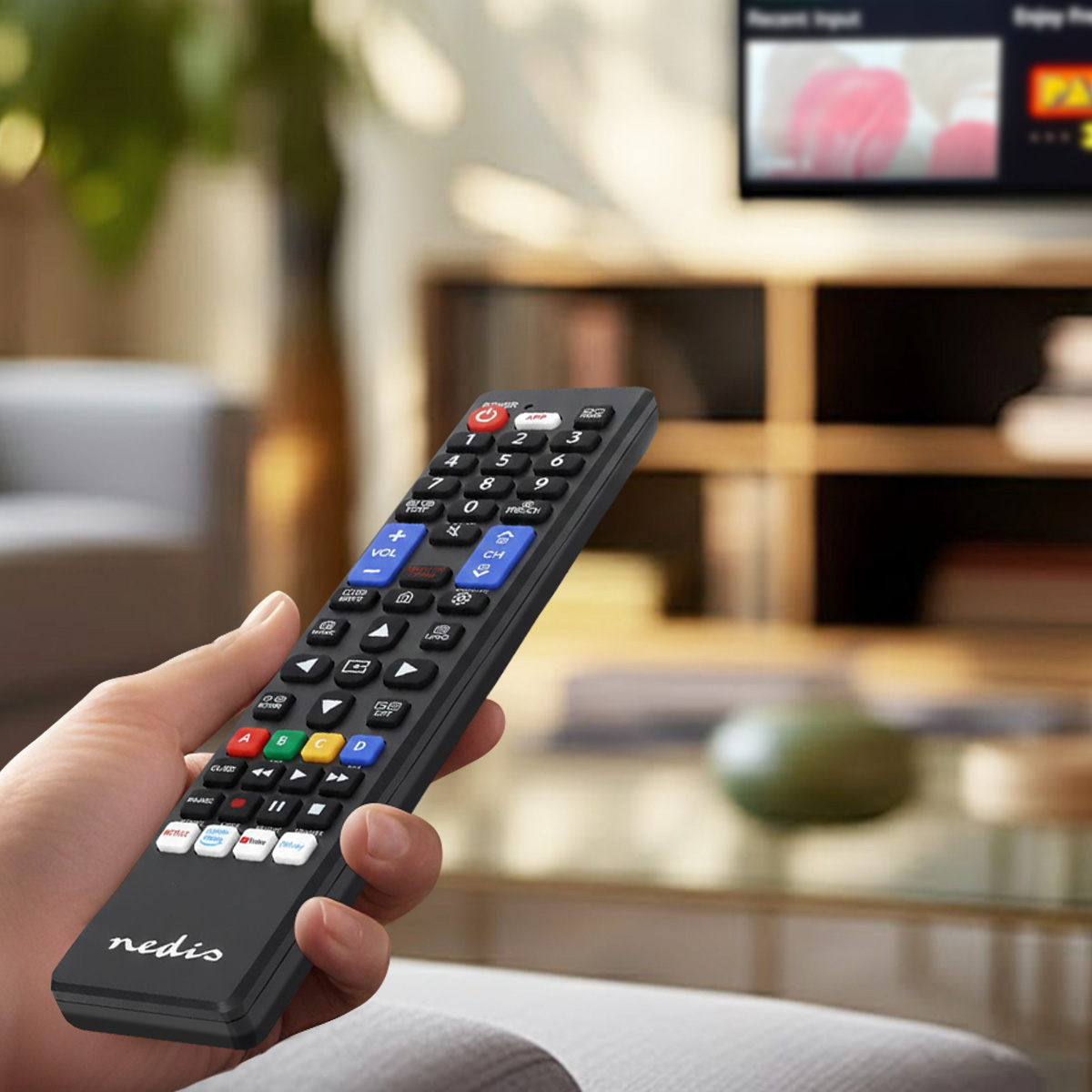 Dłoń trzyma czarny pilot Nedis przed rozmytym telewizorem i meblami.