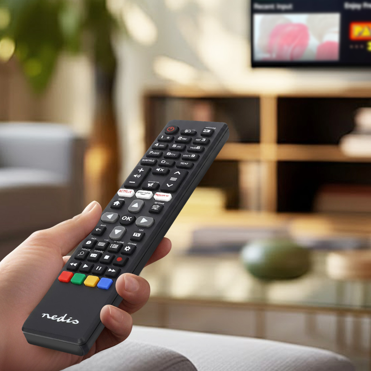 Dłoń trzyma czarny pilot z wieloma przyciskami przed rozmytym telewizorem.