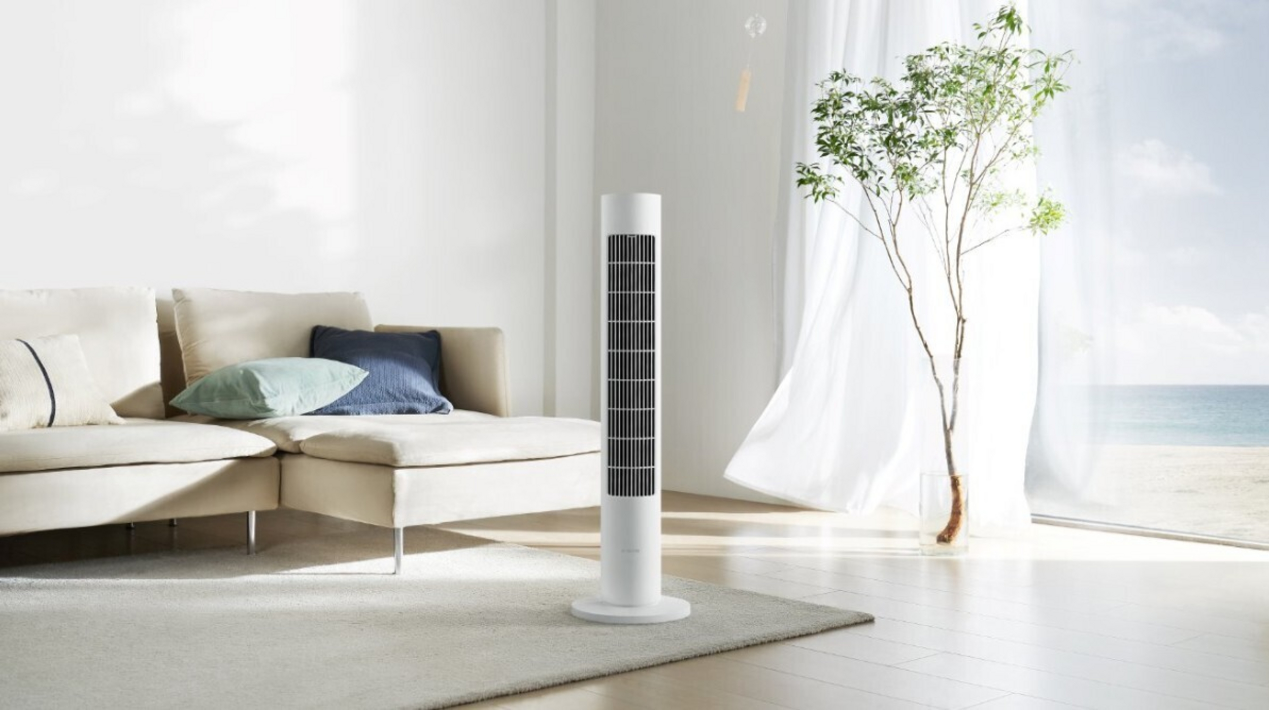 Biały wentylator wieżowy obok sofy narożnej z poduszkami. Drzewo w szkle.