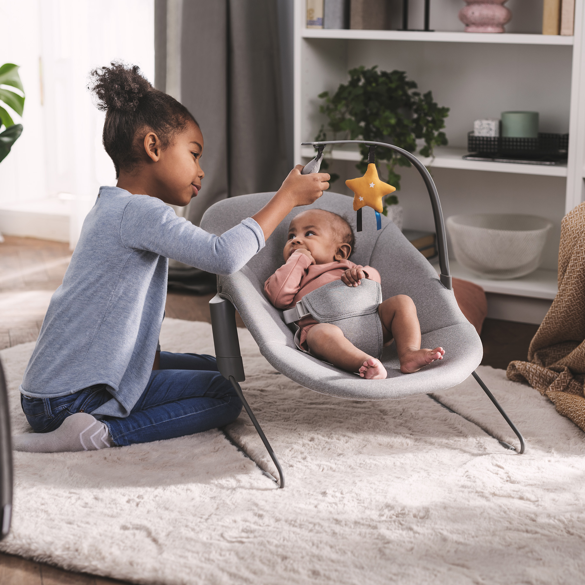 Ein älteres Kind sitzt auf dem Boden und spielt mit einem Baby in einer grauen Babywippe.