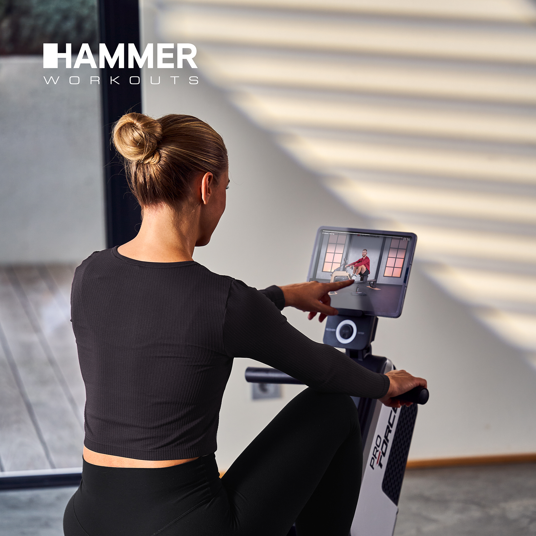 Frau auf Rudergerät schaut auf Bildschirm mit Trainer. Hammer Workouts Logo.