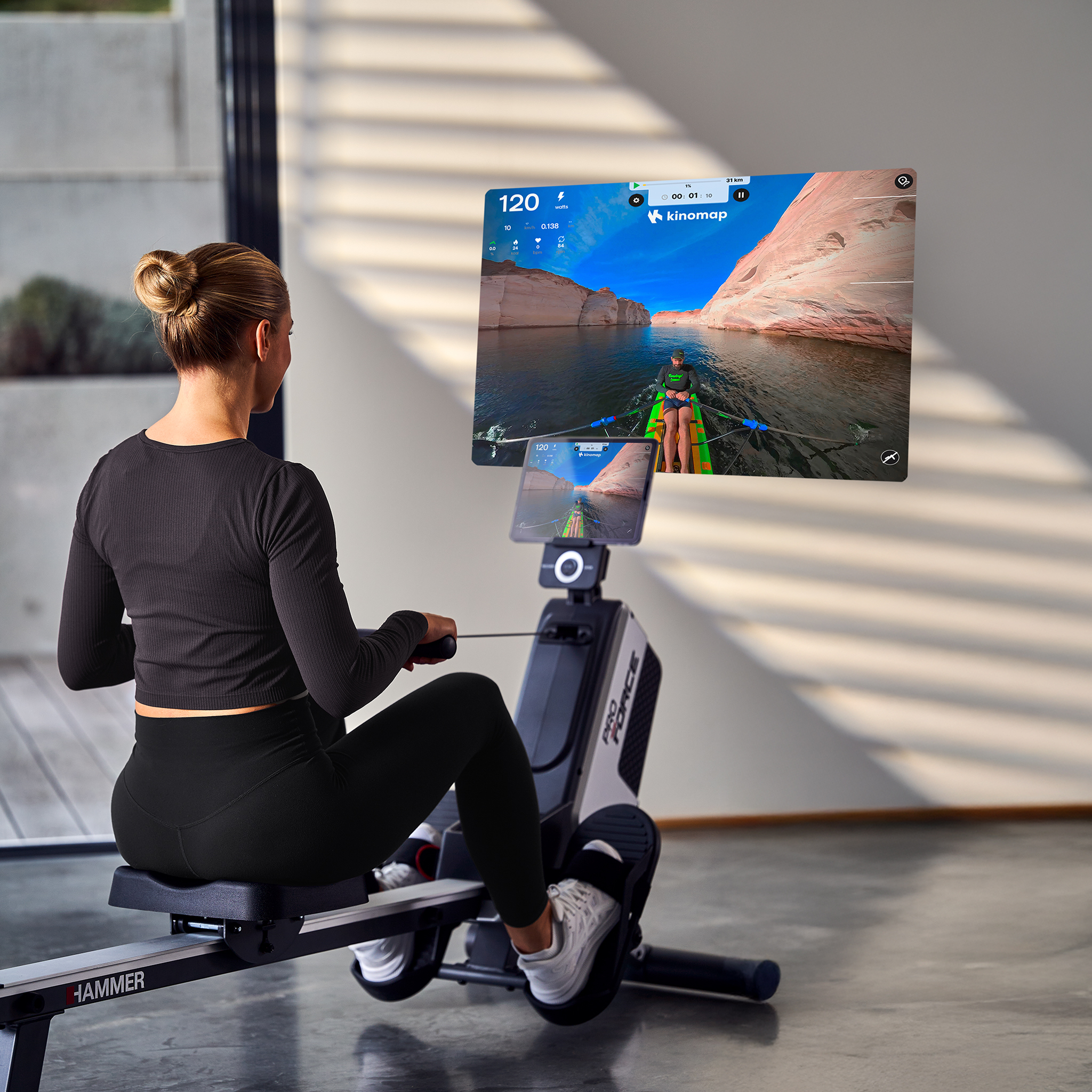 Frau rudert auf einem Ergometer vor einem Bildschirm mit einer Bootsfahrt auf einem See.