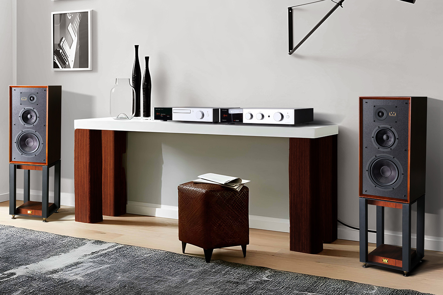 Twee luidsprekers flankeren een witte console met audioapparatuur en decoratie. Een leren poef ervoor.
