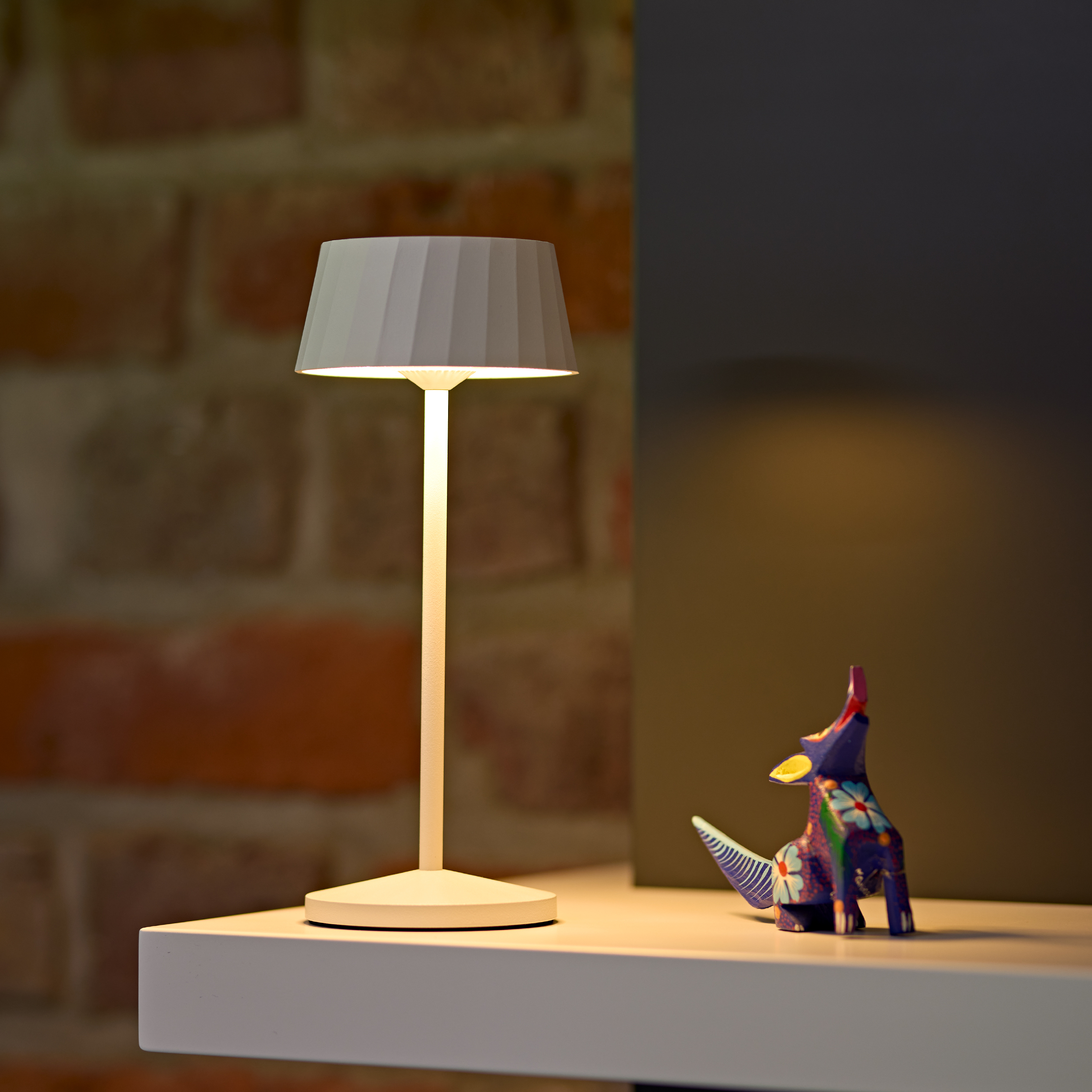Eine weiße Lampe steht neben einer bunten Tierfigur auf einem Regal.