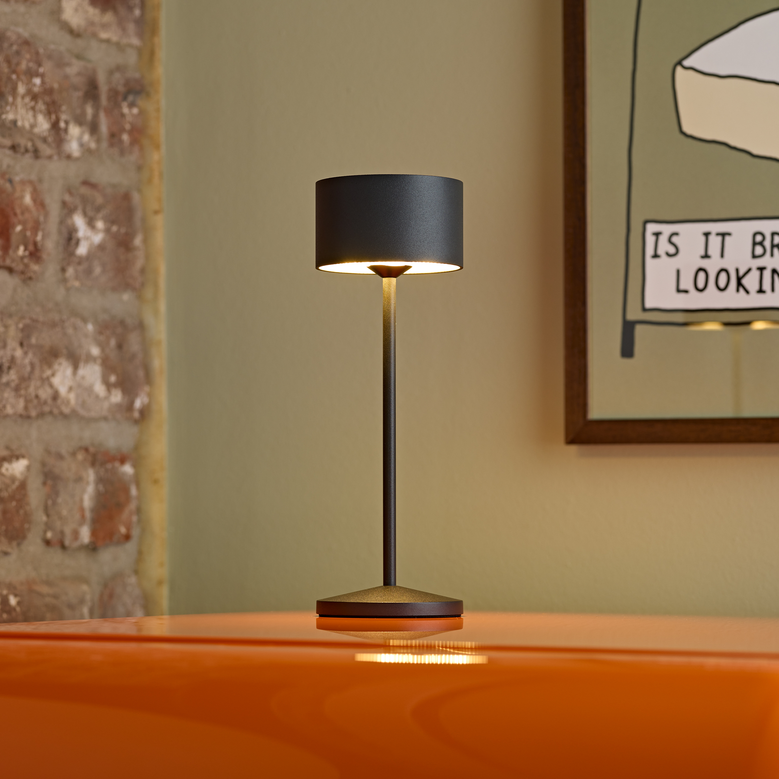 Eine dunkle Tischlampe mit warmem Licht auf einem orangen Tisch vor einer Ziegelwand.