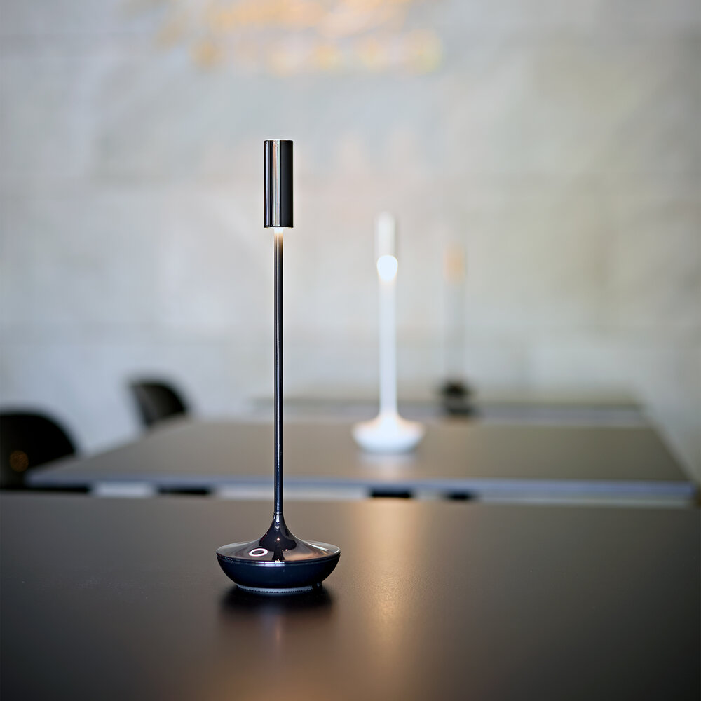 Eine schlanke, dunkle Tischlampe steht auf einem schwarzen Tisch. Im Hintergrund sind weitere Lampen.