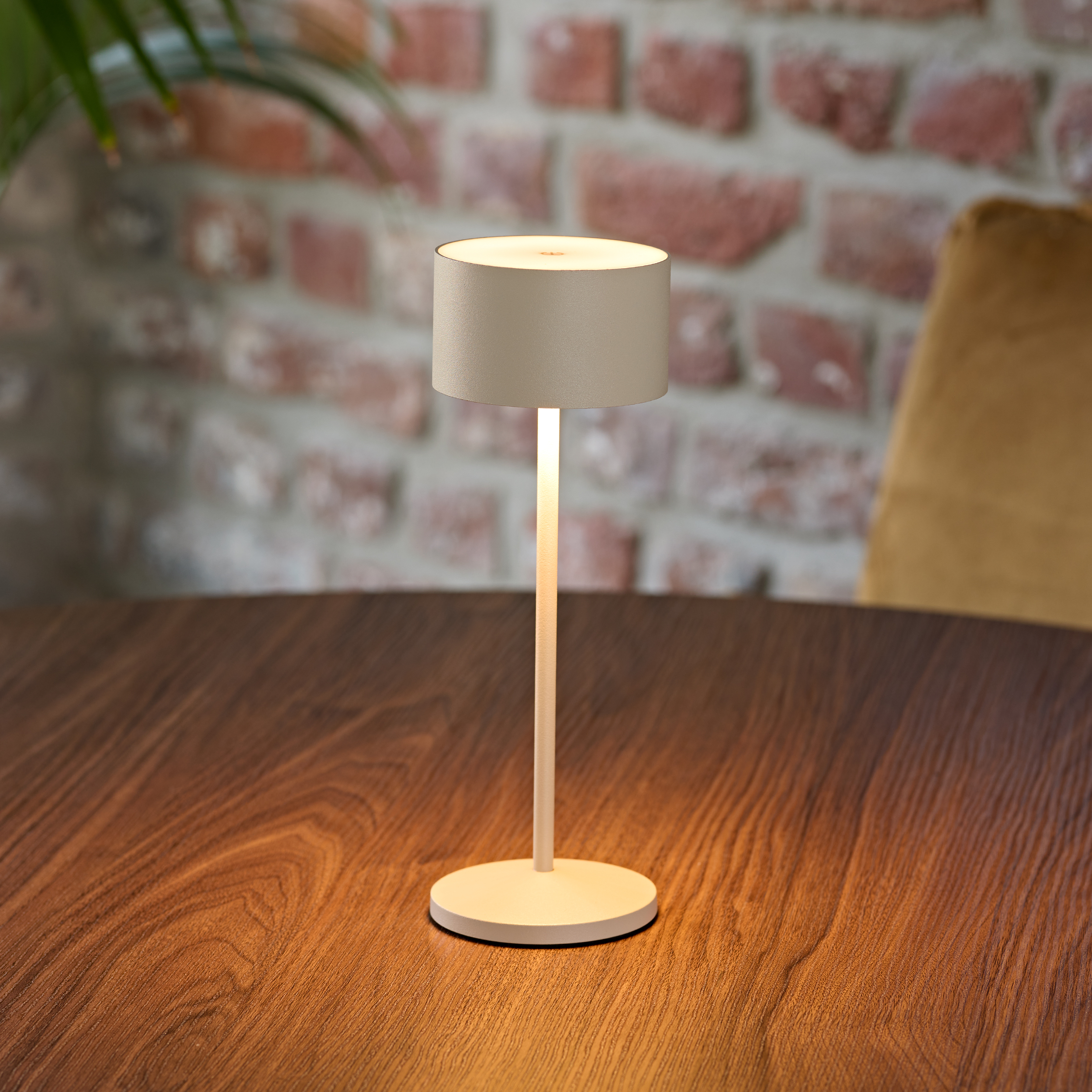 Eine helle Tischlampe steht auf einem Holztisch, Ziegelwand im Hintergrund.