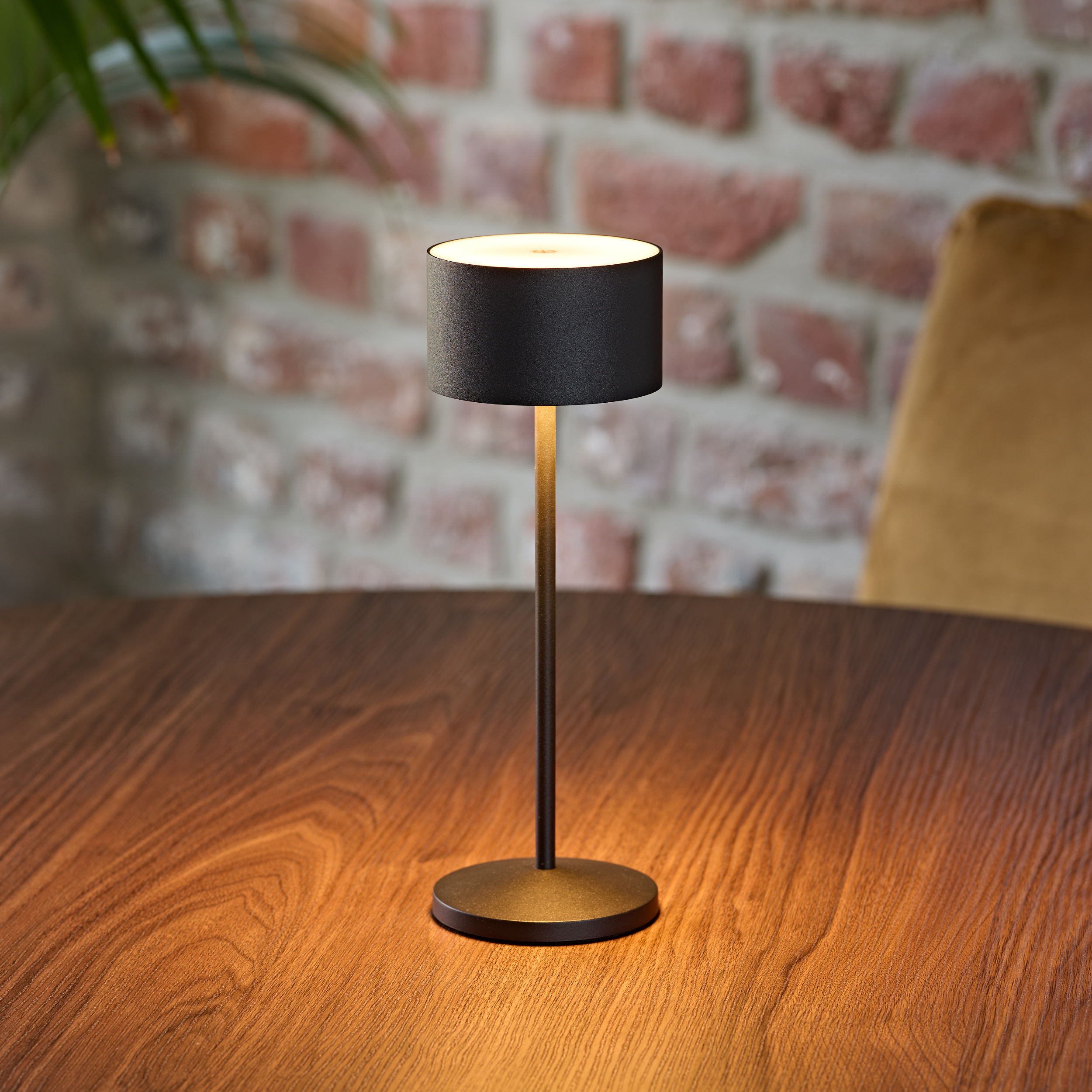 Eine schwarze Tischlampe steht auf einem Holztisch vor einer Ziegelwand.