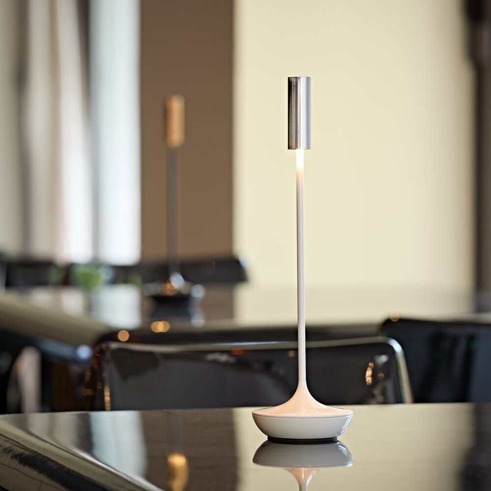 Eine schlanke, weiße Tischlampe mit silbernem Zylinder leuchtet auf einem dunklen Tisch.