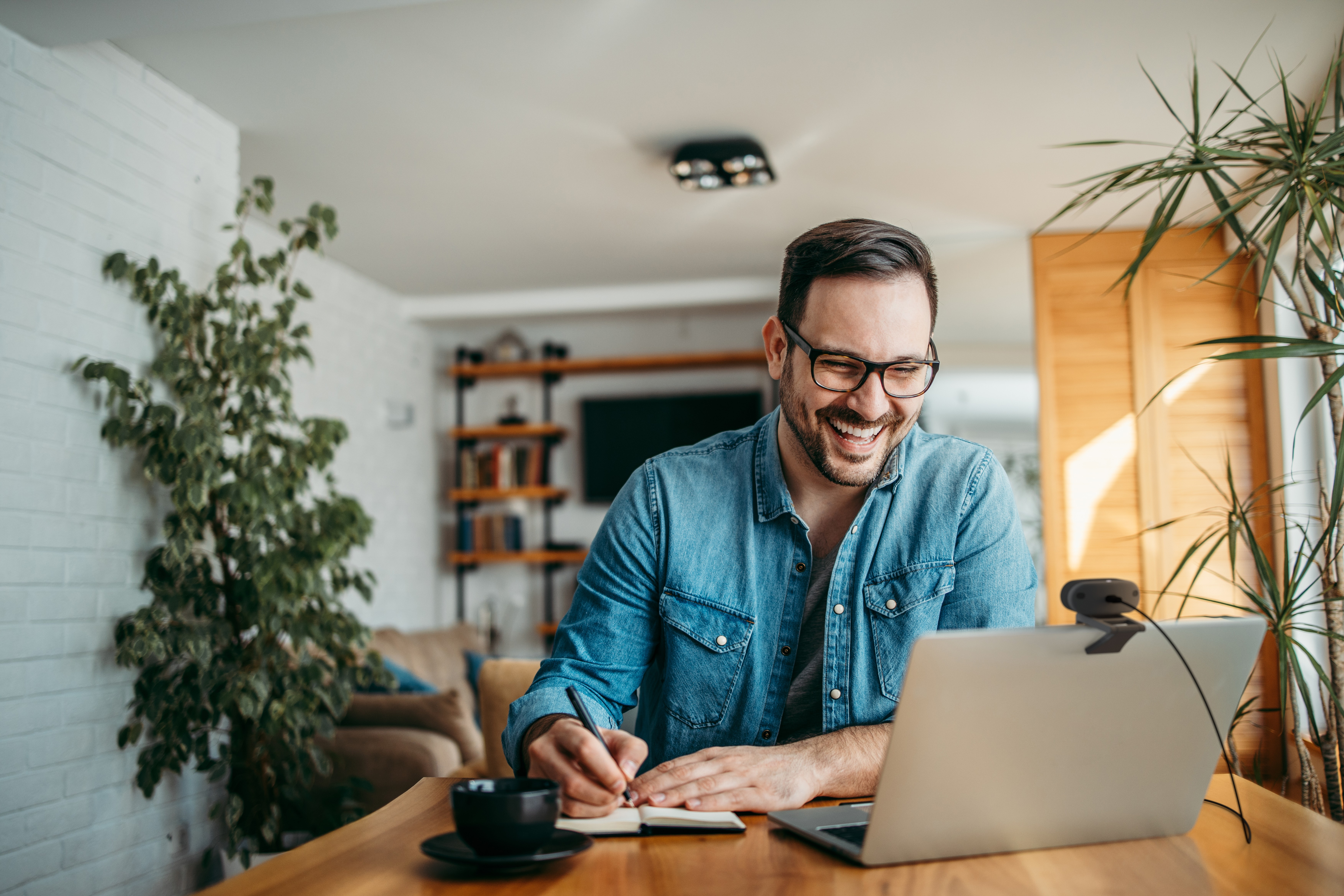 A man in glasses laughs while writing on a laptop and drinking coffee.