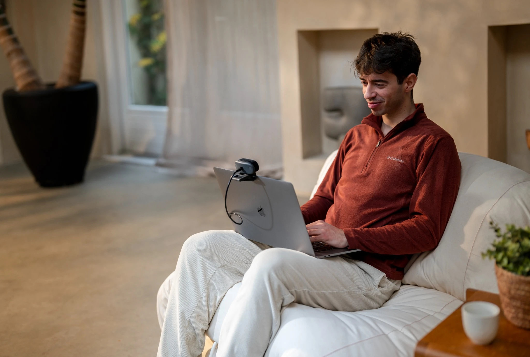 Man sits on sofa with laptop and webcam, plant in background.