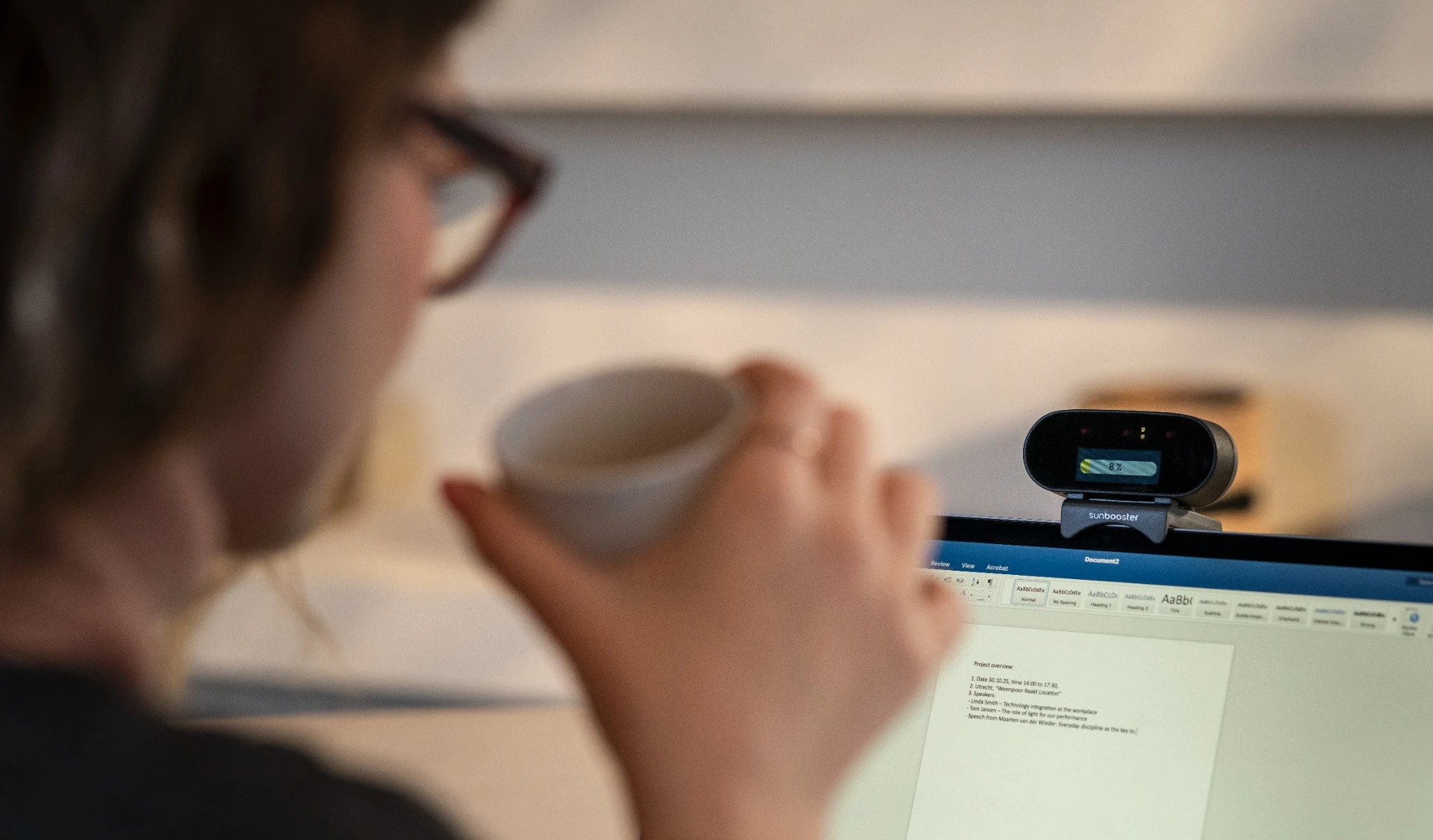 A person drinks coffee in front of a laptop with a webcam.