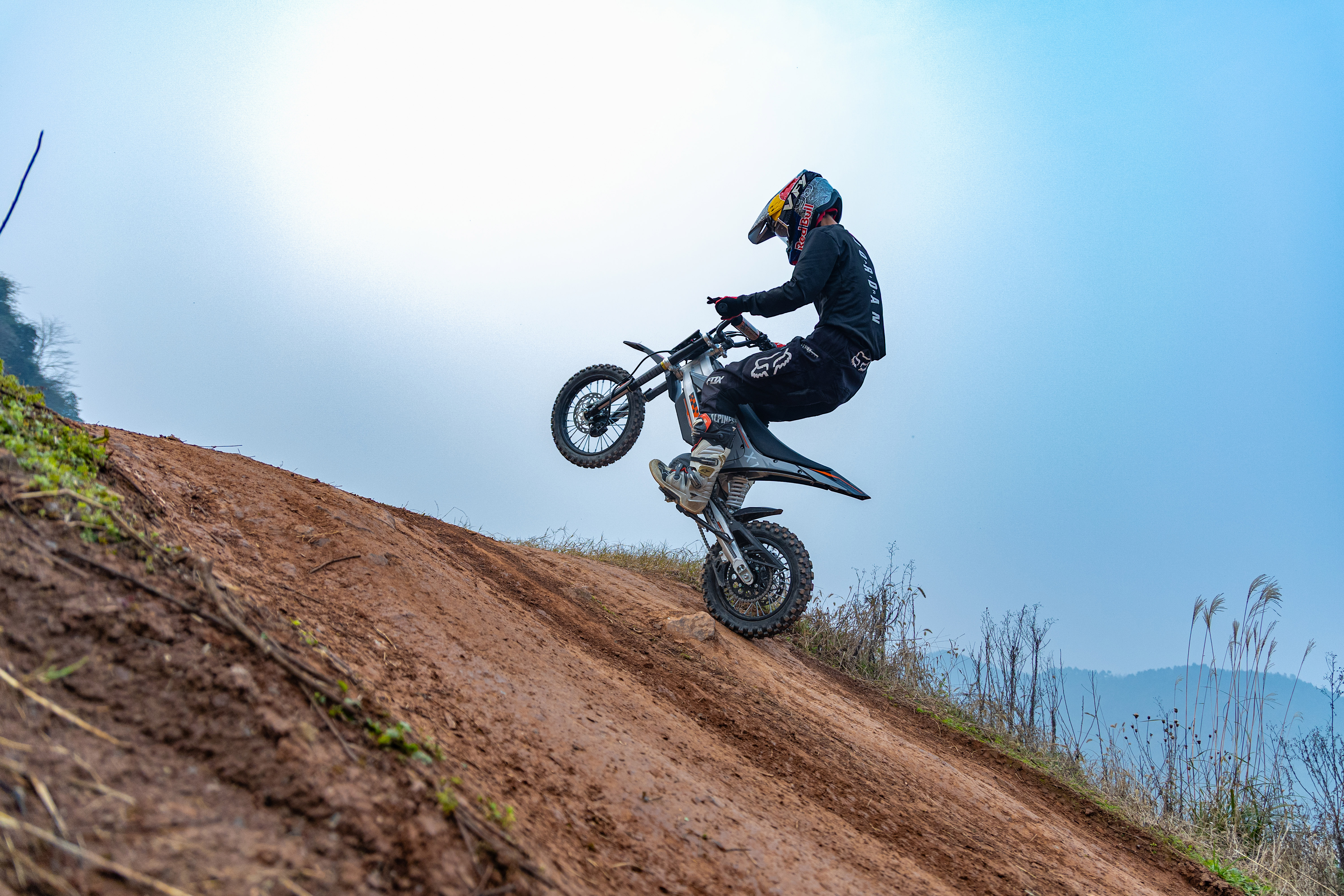 Ein Motorradfahrer macht einen Wheelie auf einer Schlammrampe unter blauem Himmel.