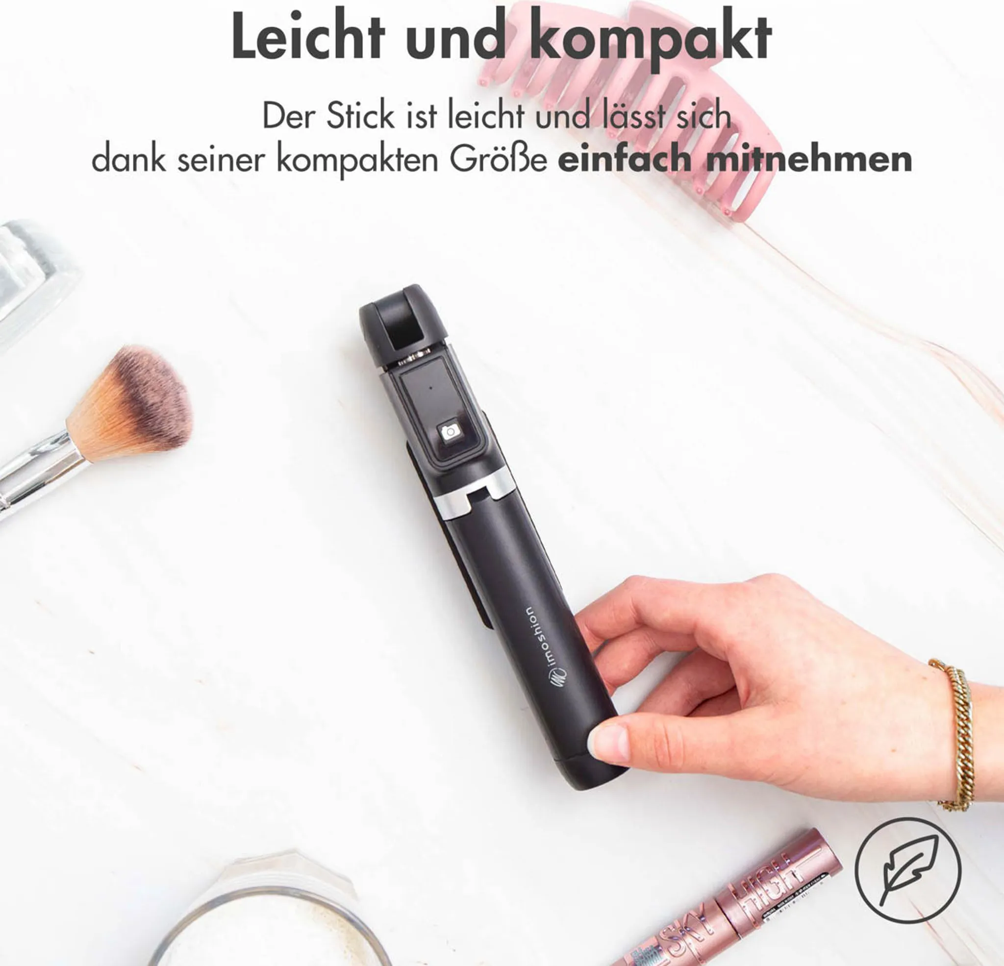Eine Hand hält einen schwarzen Selfie-Stick, neben Pinsel und Mascara.