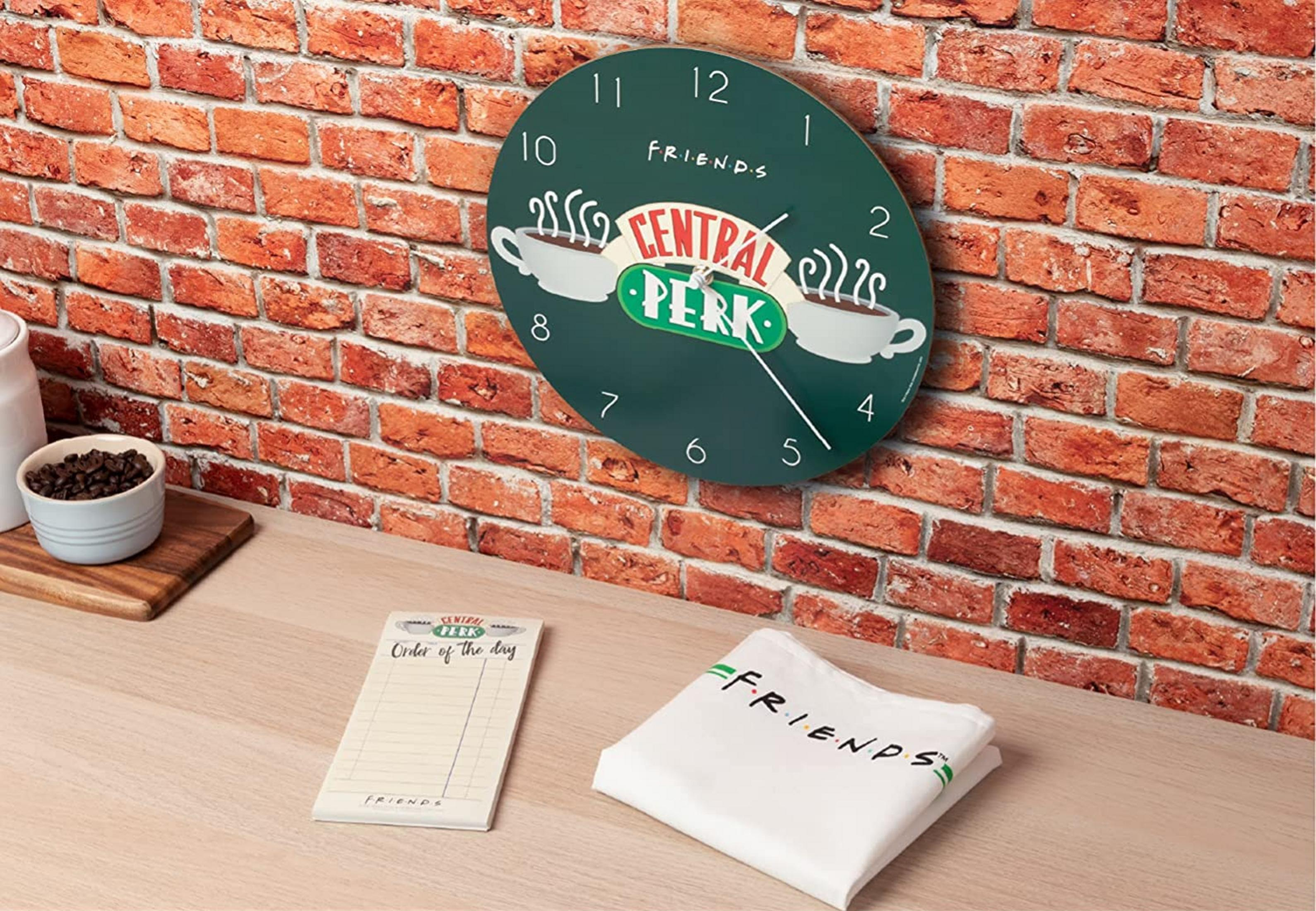 Eine Wanduhr mit dem Central Perk-Logo hängt an einer Ziegelwand. Daneben liegen Notizblöcke und ein gefaltetes T-Shirt.