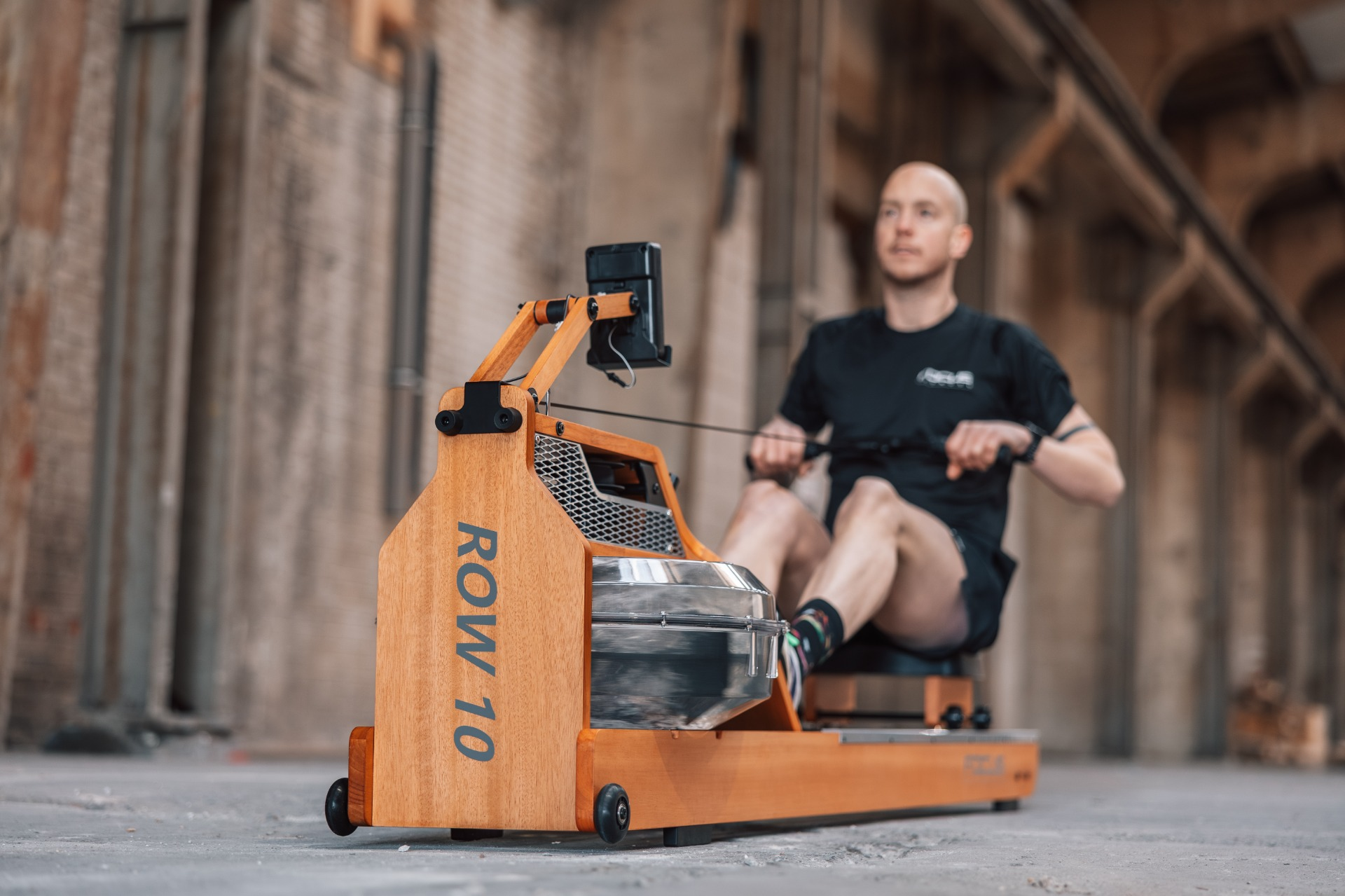 A man rows on a wooden rowing machine inside an industrial building.