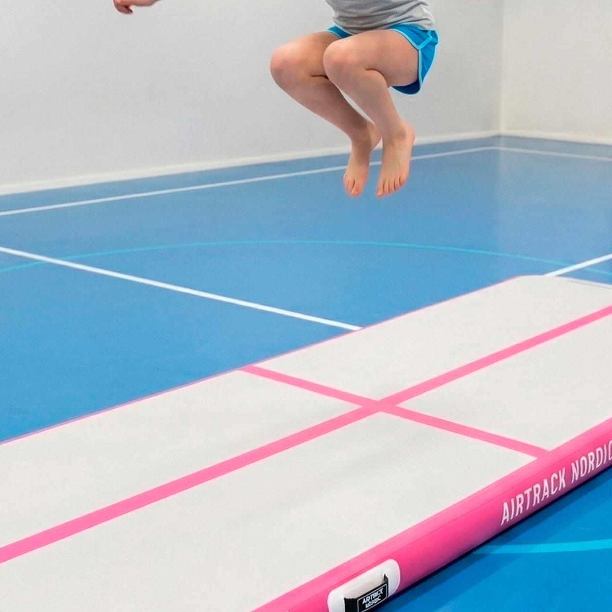 Dziecko w niebieskich szortach skacze boso na biało-różowej ścieżce powietrznej w niebieskiej sali gimnastycznej z białymi liniami.