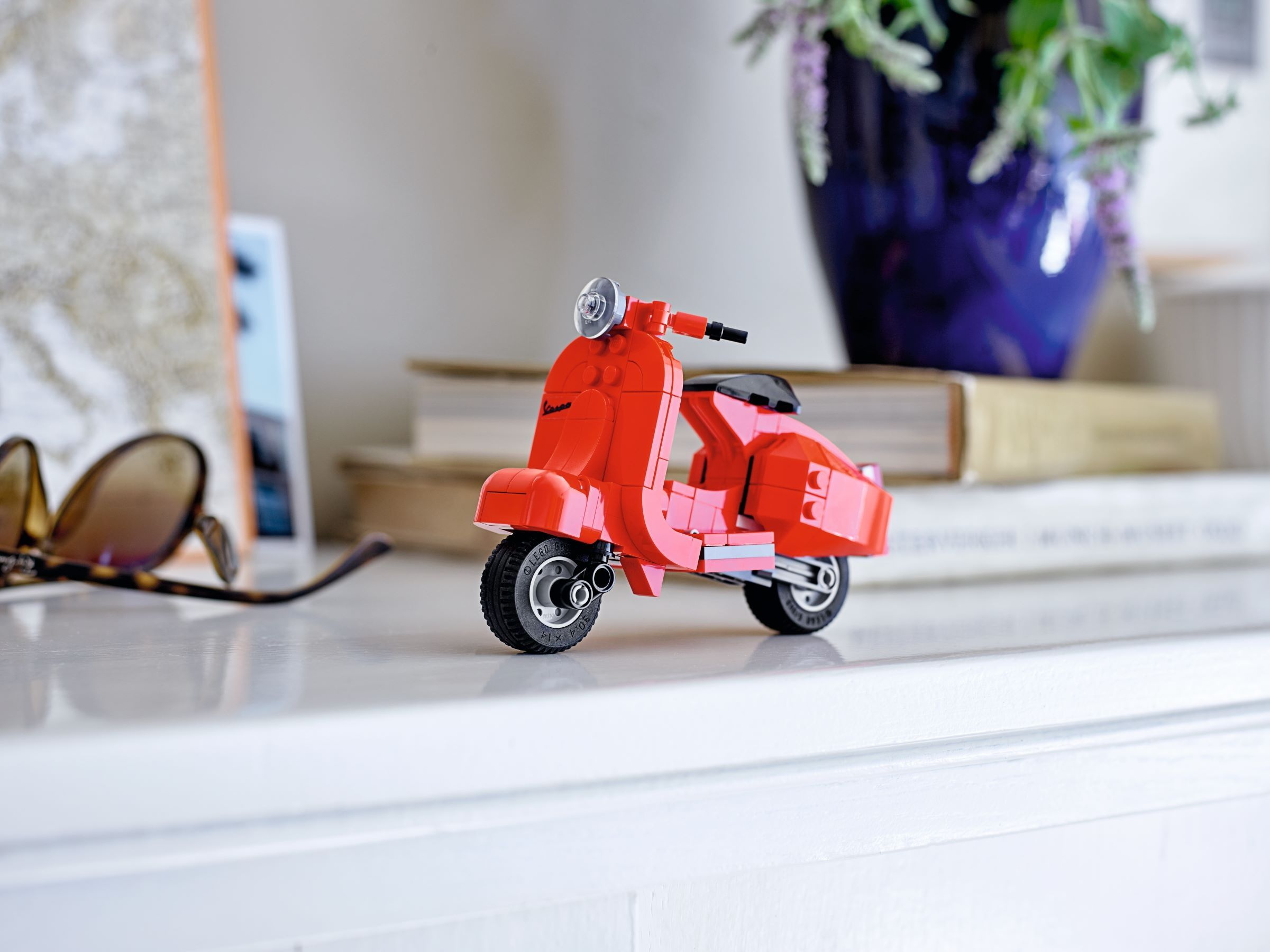 Uno scooter Lego rosso siede su uno scaffale vicino a libri e un vaso.