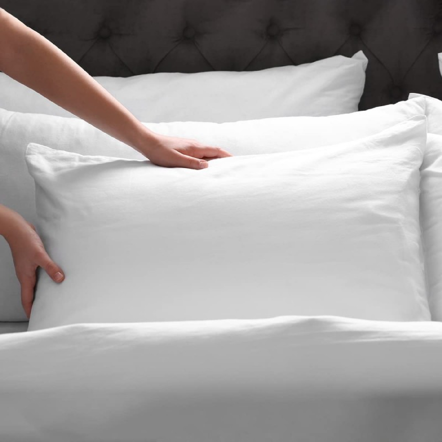 Una mano coloca una almohada blanca sobre una cama con cabecero oscuro.