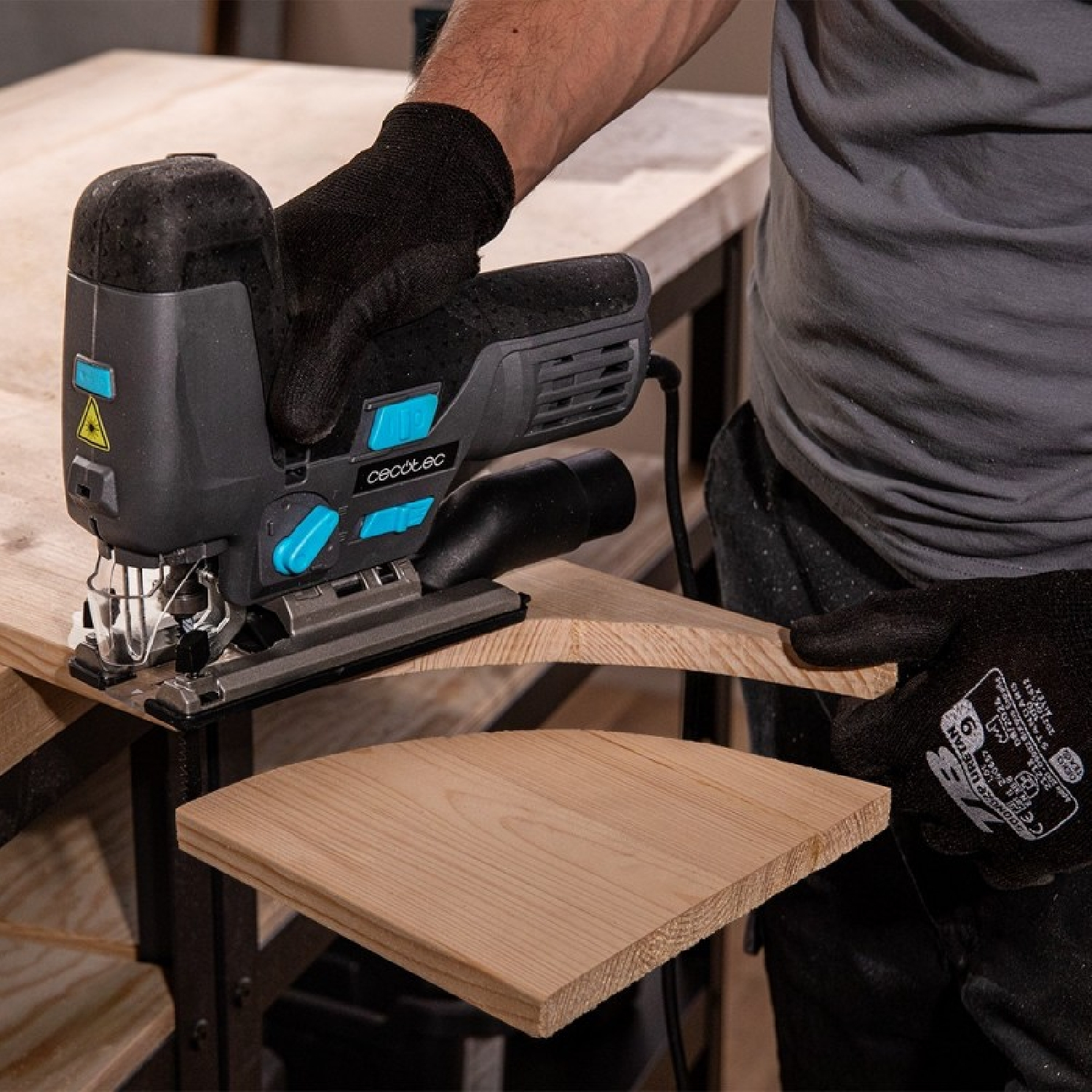 Una persona con guantes negros usa una sierra de calar para cortar una pieza curva de madera en un banco de trabajo.