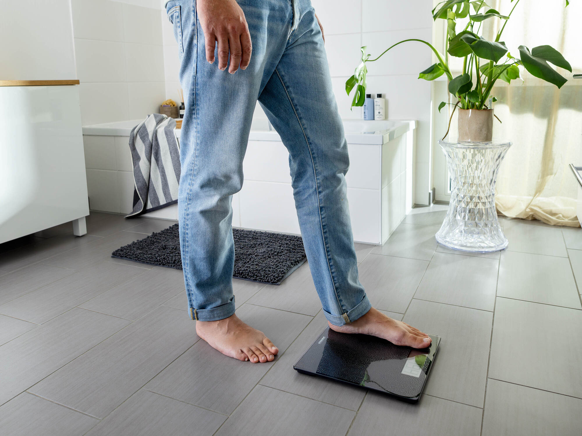 Eine Person in blauen Jeans steht barfuß in einem Badezimmer, ein Fuß auf einer schwarzen Digitalwaage, mit einer Pflanze im Hintergrund.