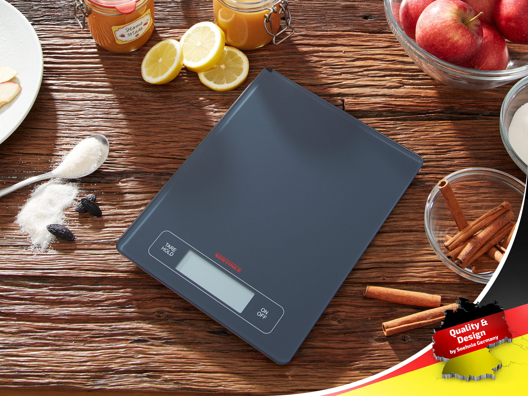 Szara waga kuchenna leży na drewnianym stole z dodatkami, takimi jak jabłka, cytryny, cukier i cynamon.