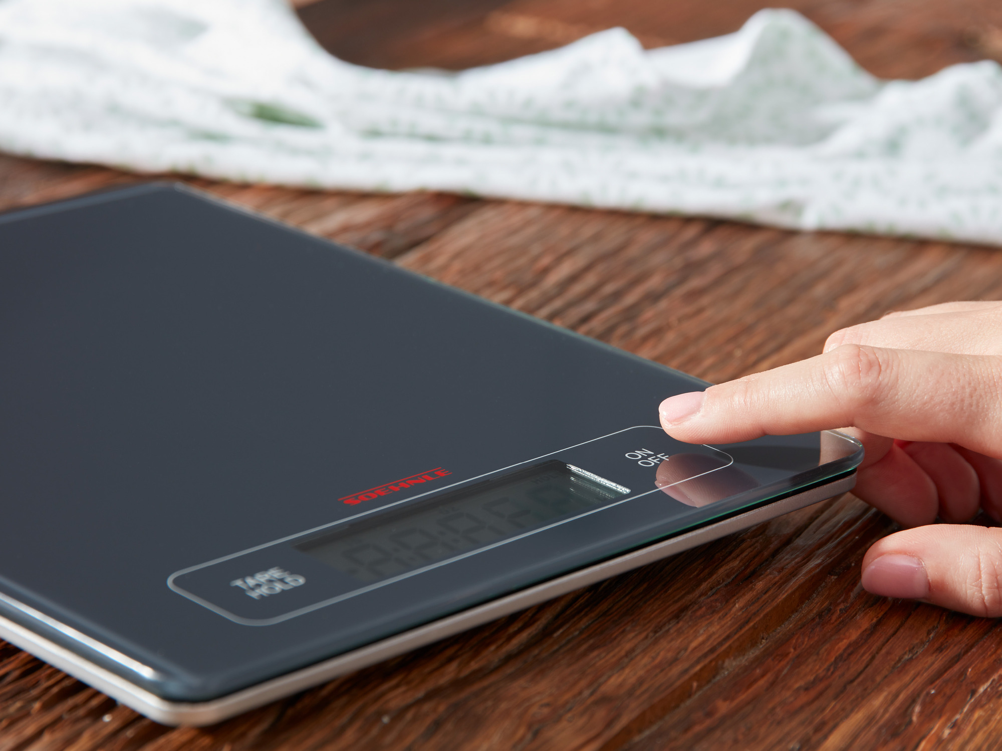 Dłoń naciska przycisk włączania/wyłączania ciemnej wagi kuchennej na drewnianym stole.