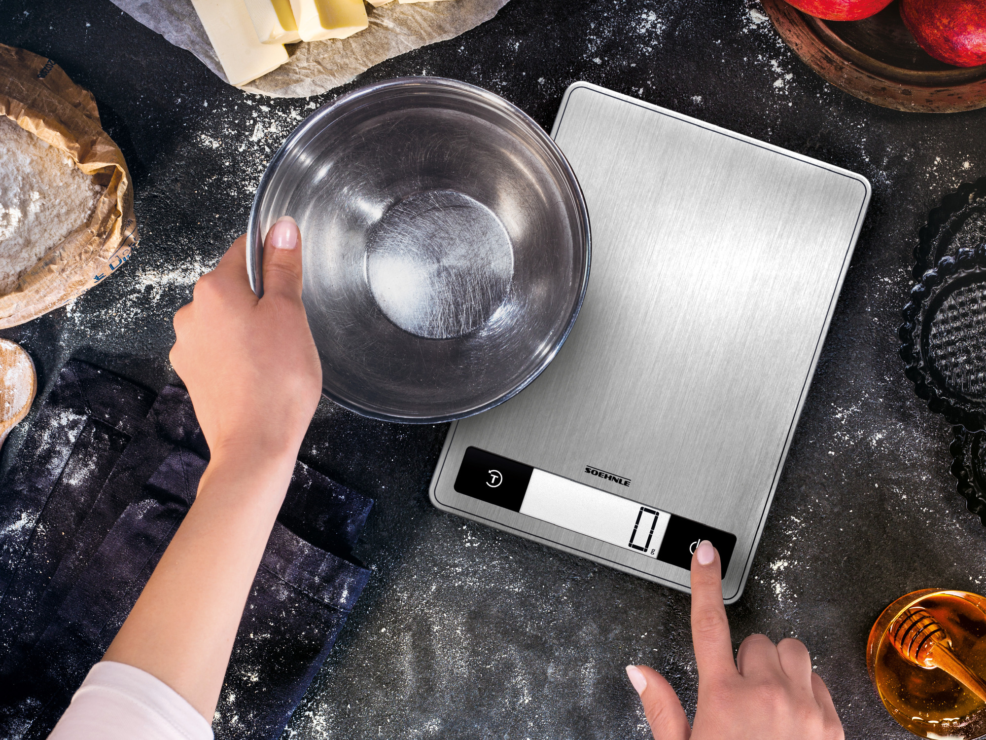 Ręce używają wagi kuchennej z miską, obok mąki, masła i jabłek.