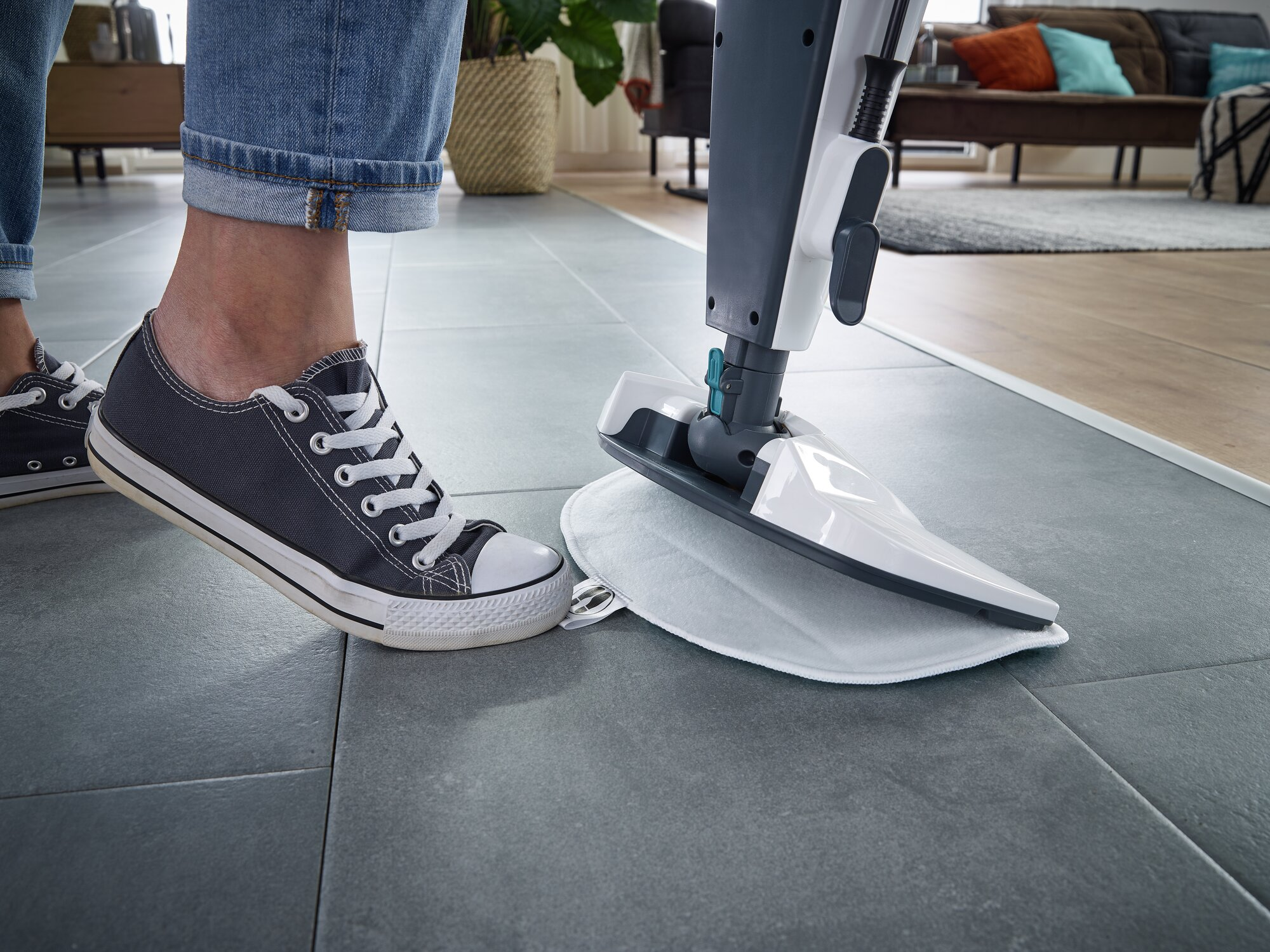 Eine Person in Jeans und Turnschuhen benutzt einen Dampfreiniger auf grauem Fliesenboden.
