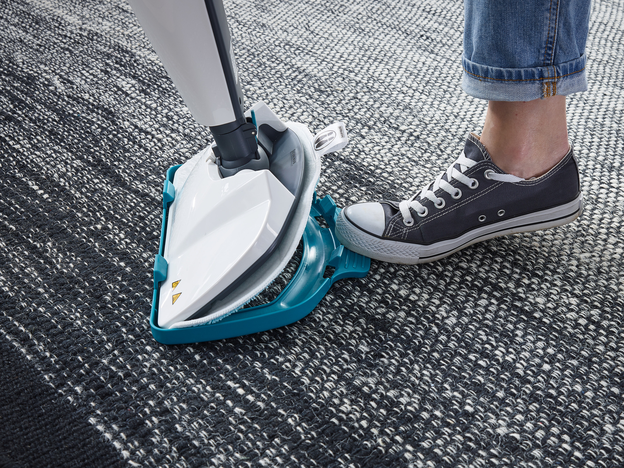 Ein Dampfreinigerkopf steht auf einem dunklen Teppich neben einem Fuß im Turnschuh.