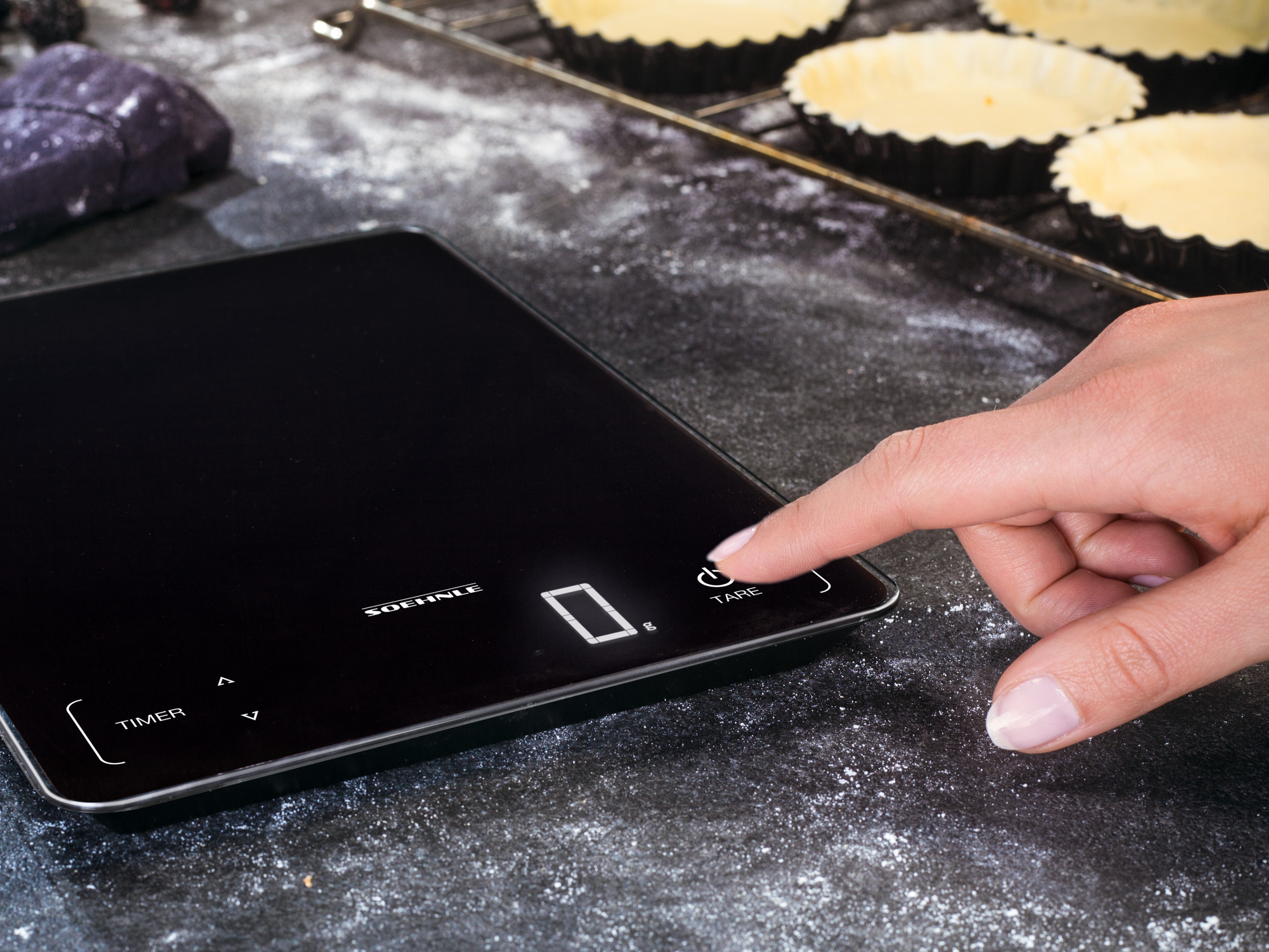 Dłoń naciska przycisk zasilania wagi kuchennej, obok ciasta i foremek do tart.