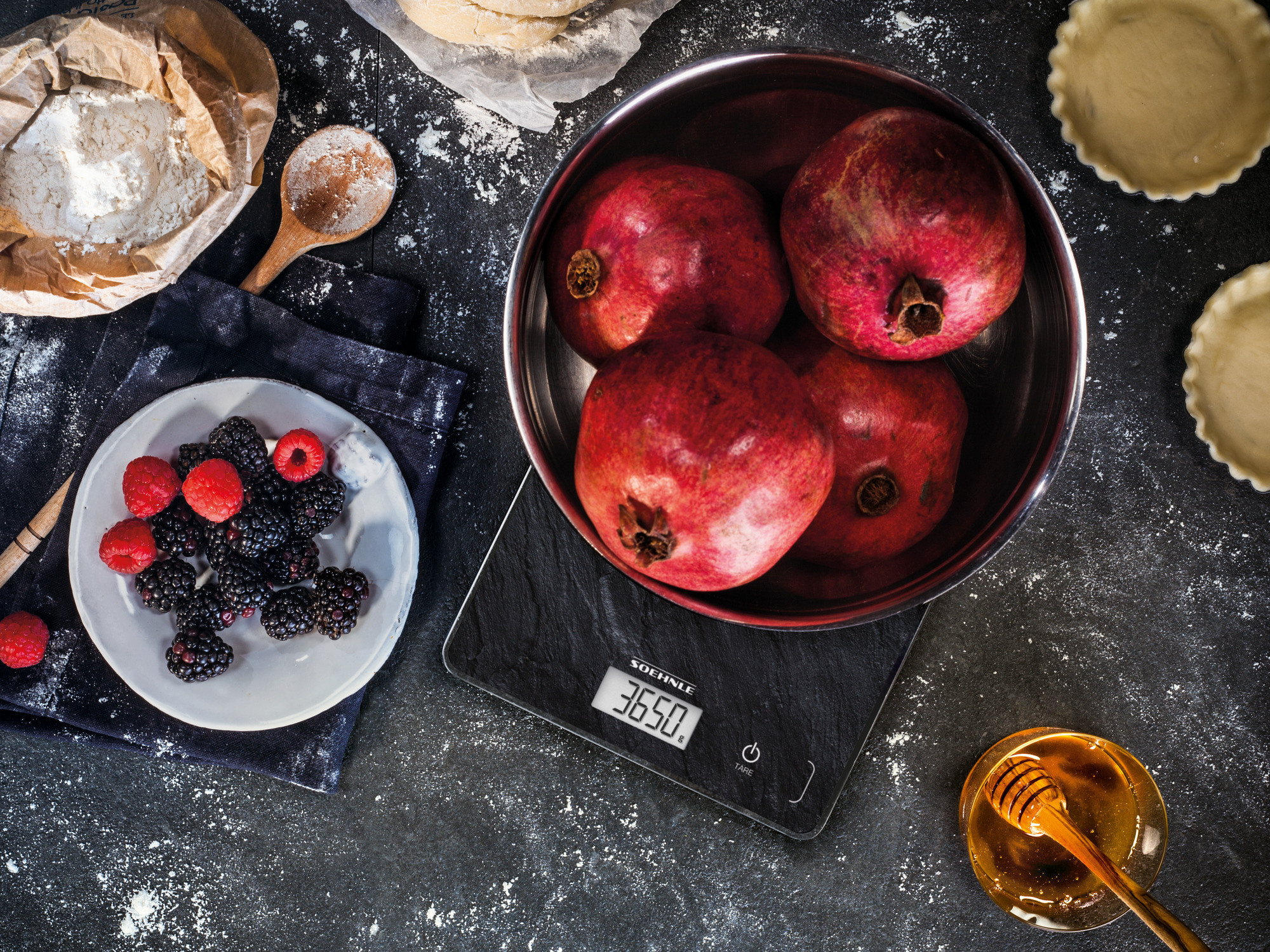 Granaty, jagody, miód, waga i składniki do pieczenia na ciemnej powierzchni.