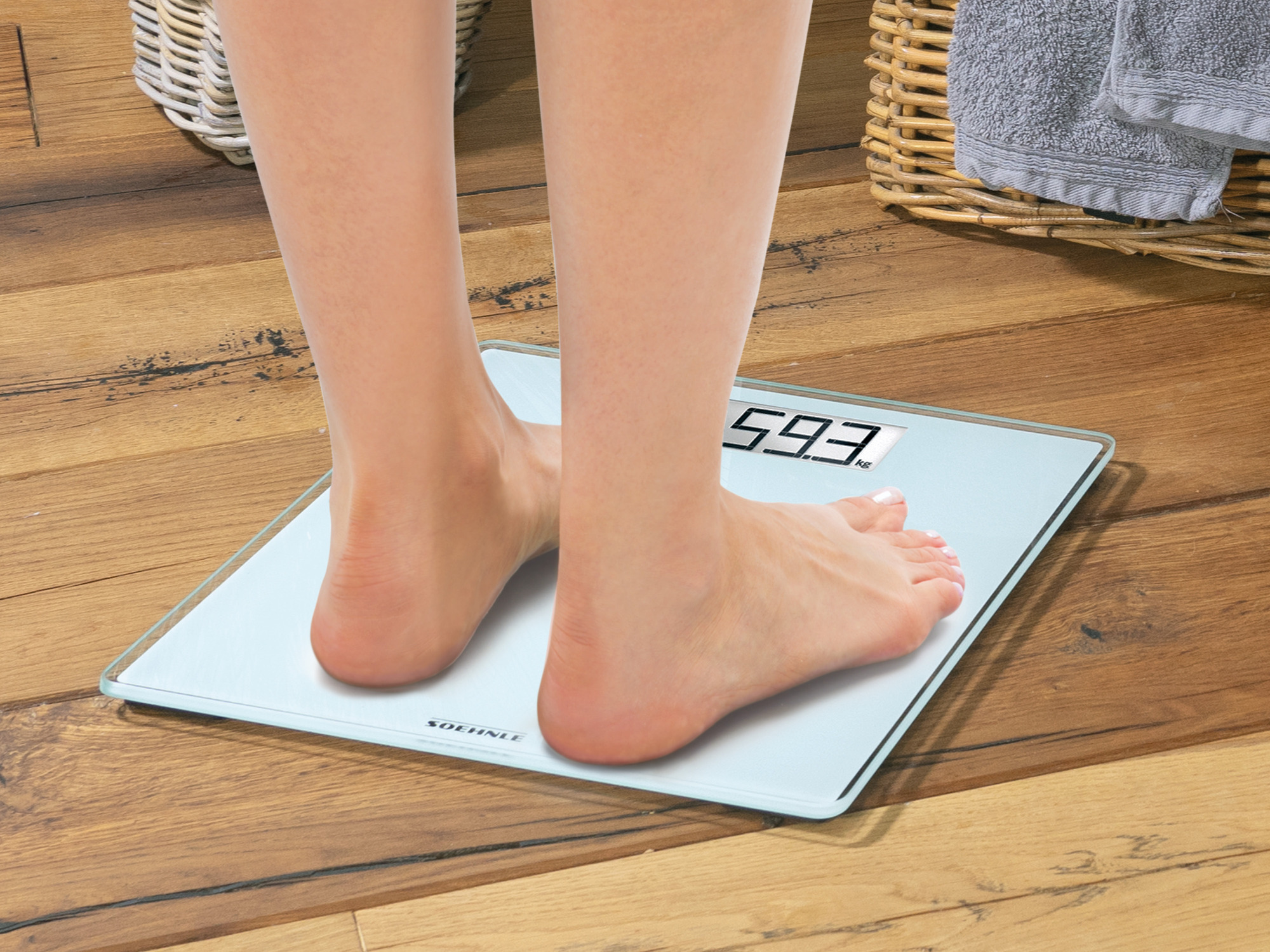 Stopy na wadze pokazującej 59.3 KG. Drewniana podłoga i kosze w tle.