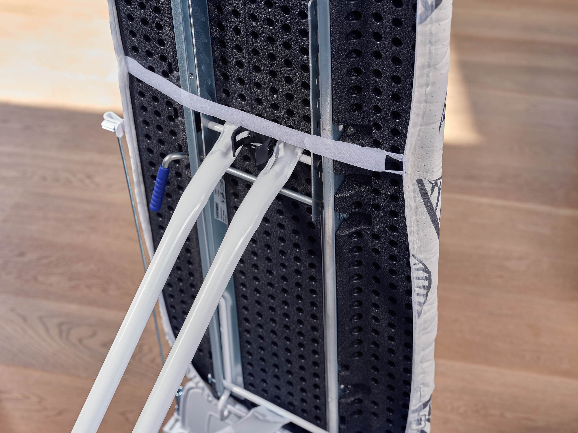 Zbliżenie stelaża deski do prasowania z białymi rurkami i niebieskim uchwytem.