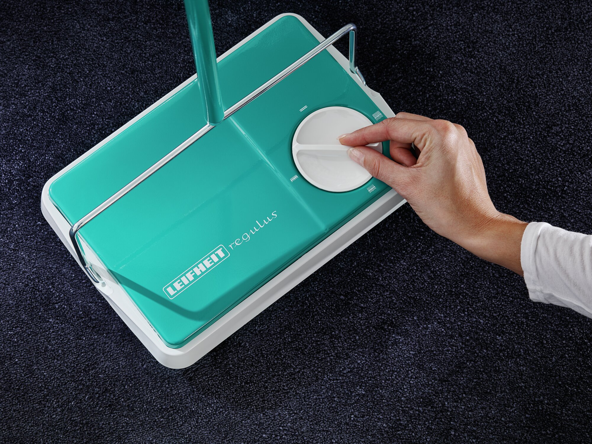 Hand dreht Knopf an türkisfarbener Bodenbürste auf dunklem Teppich.