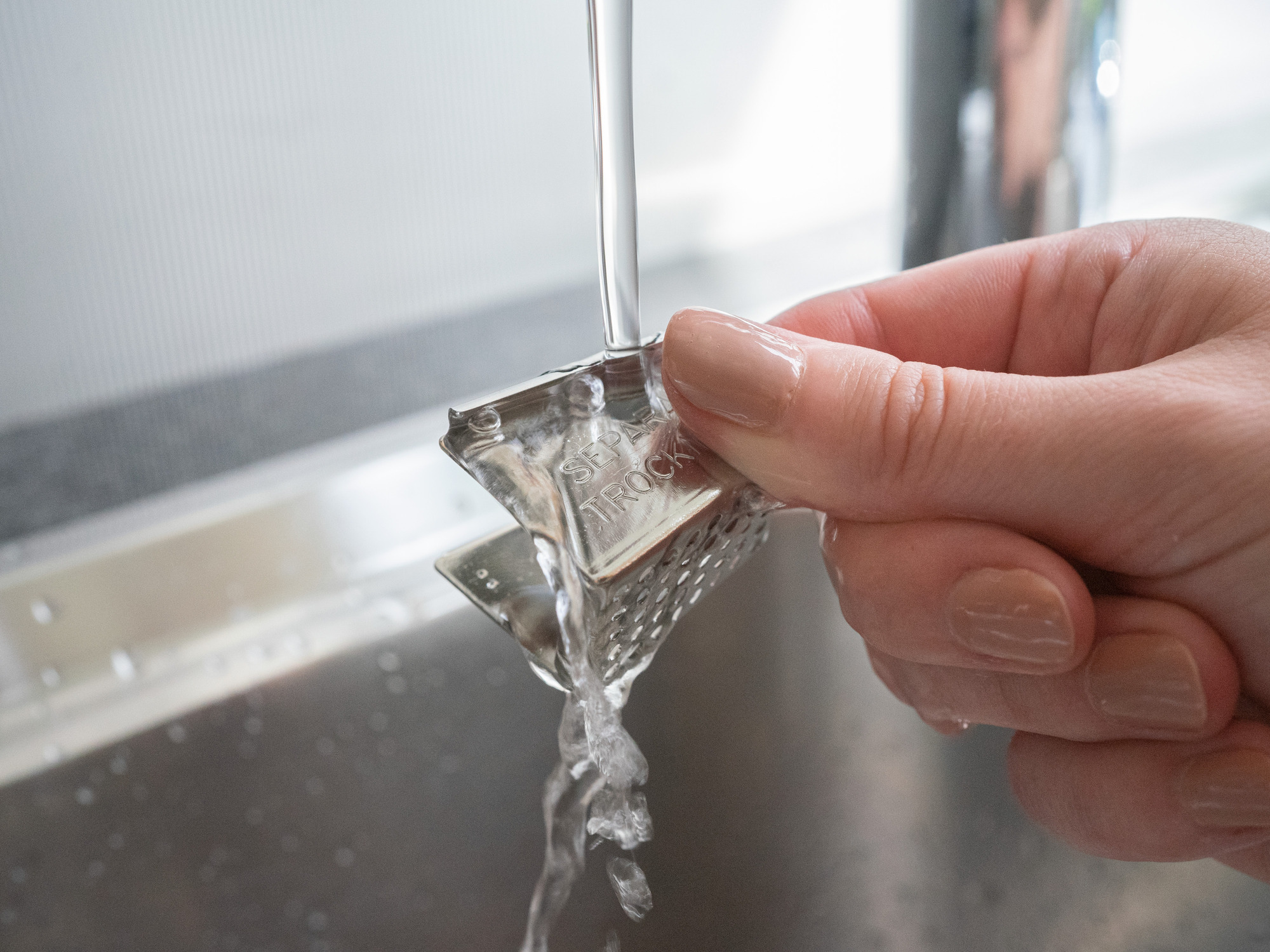 Eine Hand hält ein kleines Metallobjekt unter fließendem Wasser in einer Spüle.