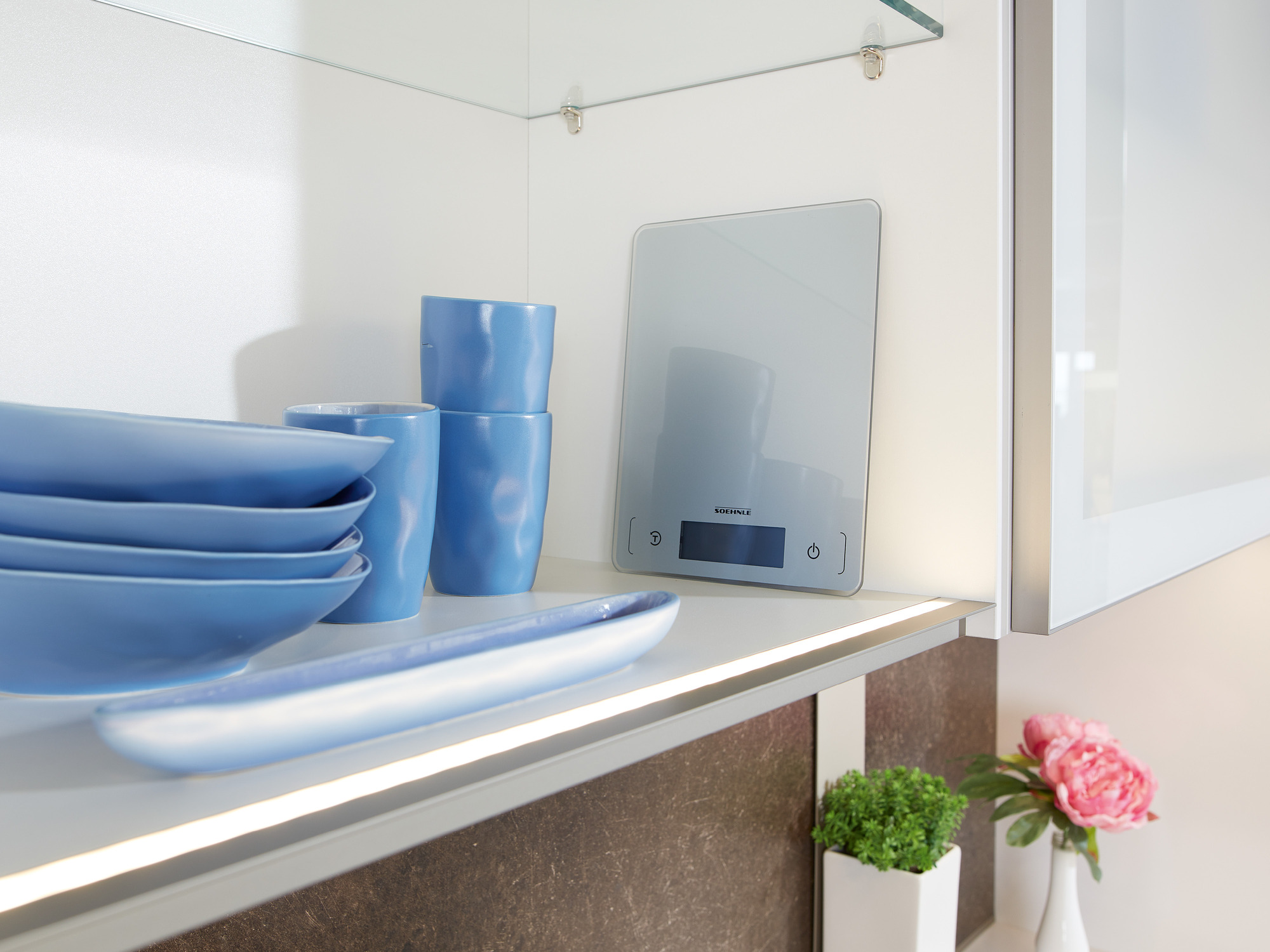 Niebieskie talerze, kubki, waga i kwiaty na półce.