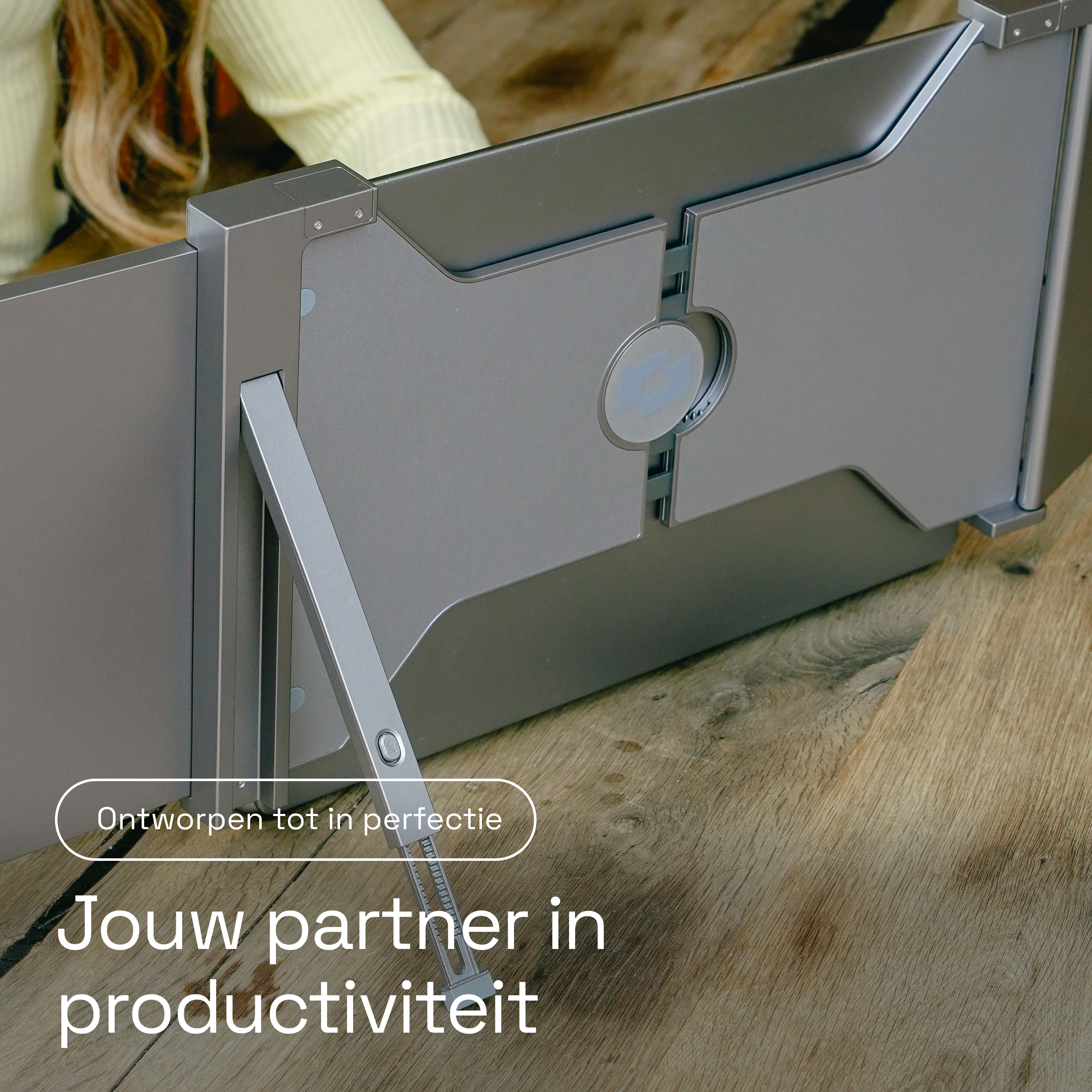 Een zilveren laptopstandaard op een houten tafel met een persoon op de achtergrond.