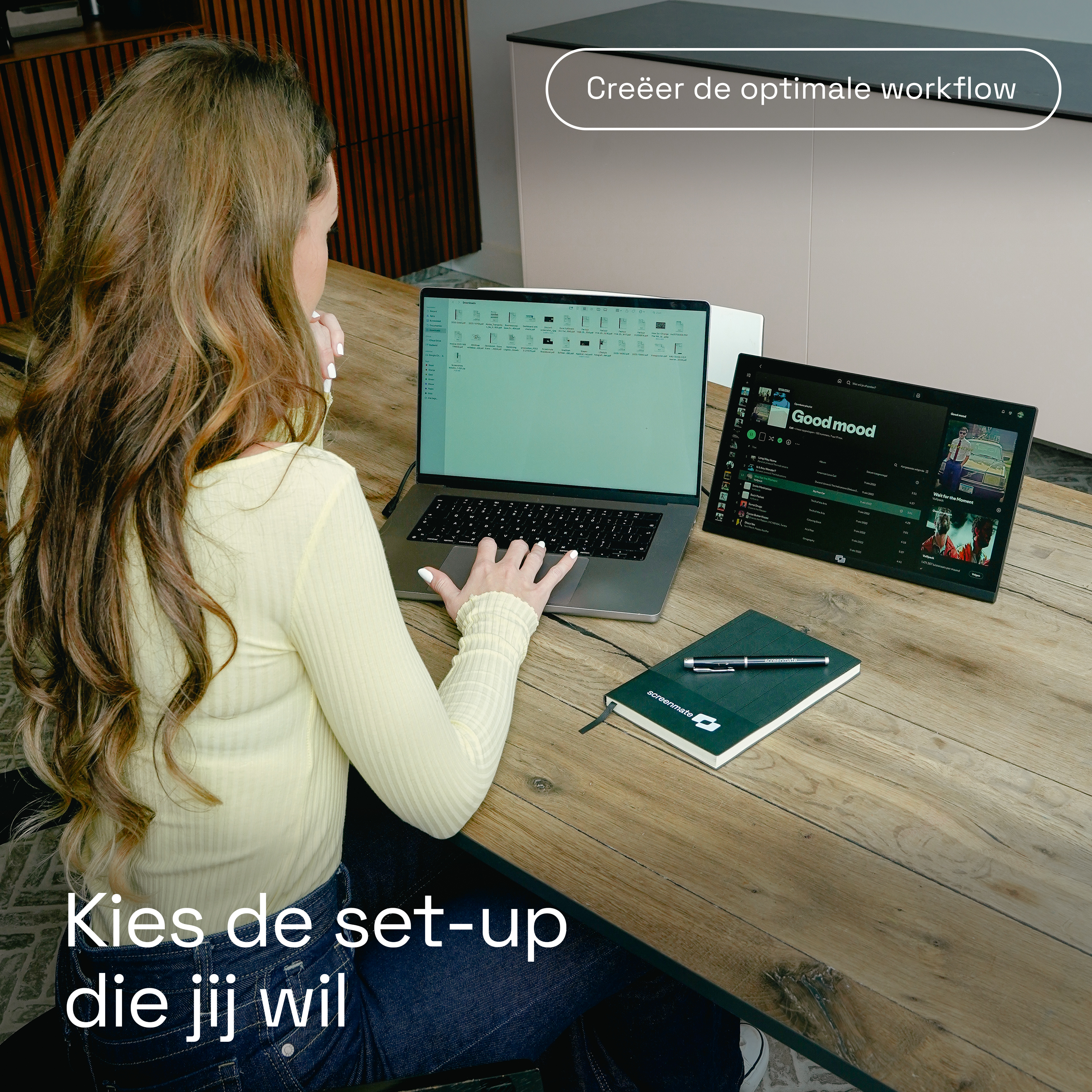 Een vrouw aan een houten tafel met laptop en tablet. Een notitieboek ligt ernaast.