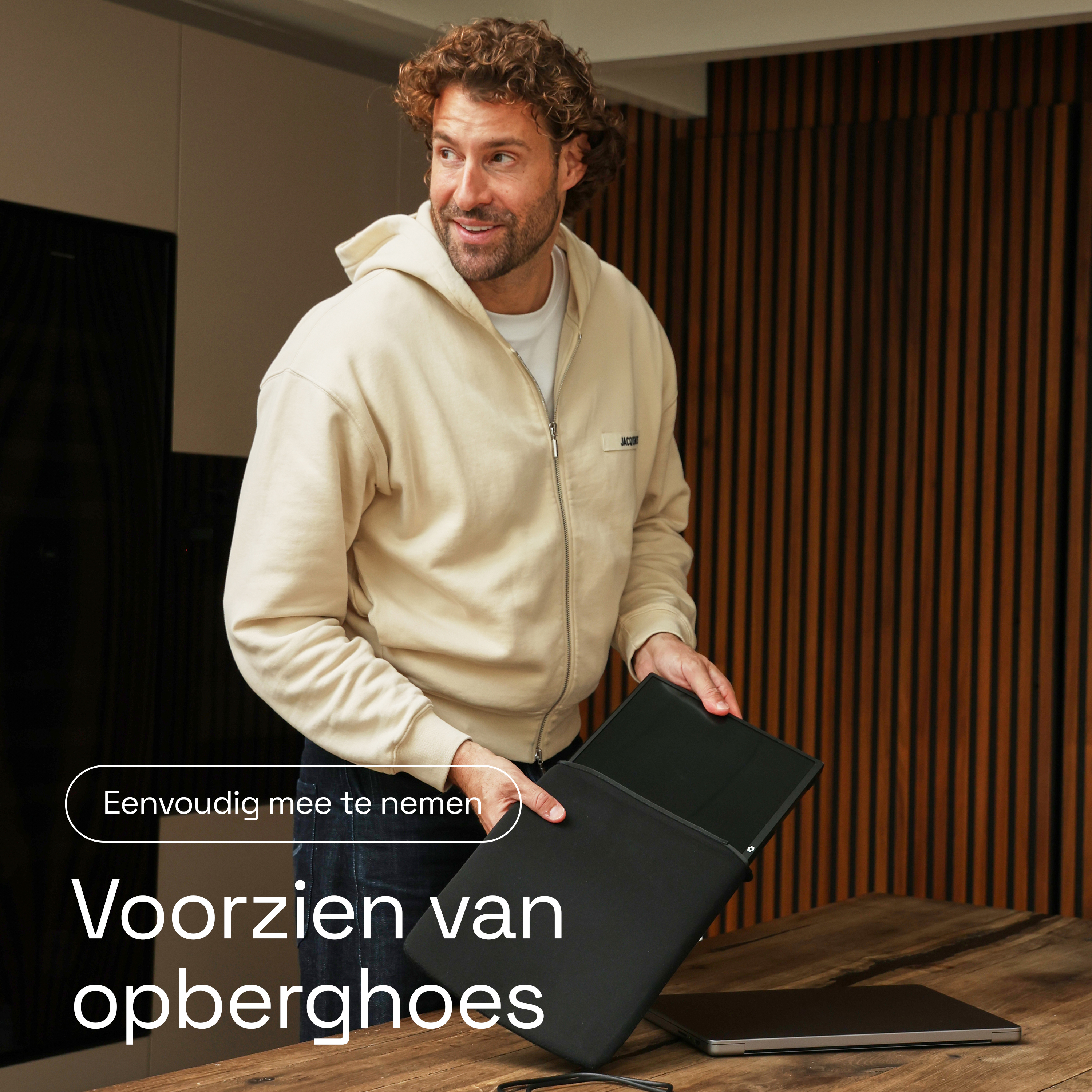 Man met krullend haar houdt zwarte laptoptas op houten tafel.