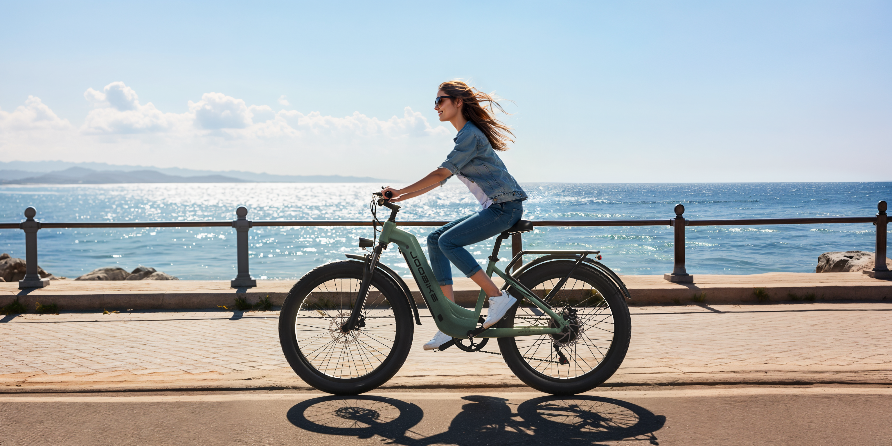 Una mujer monta una e-bike verde junto al mar, con montañas al fondo.