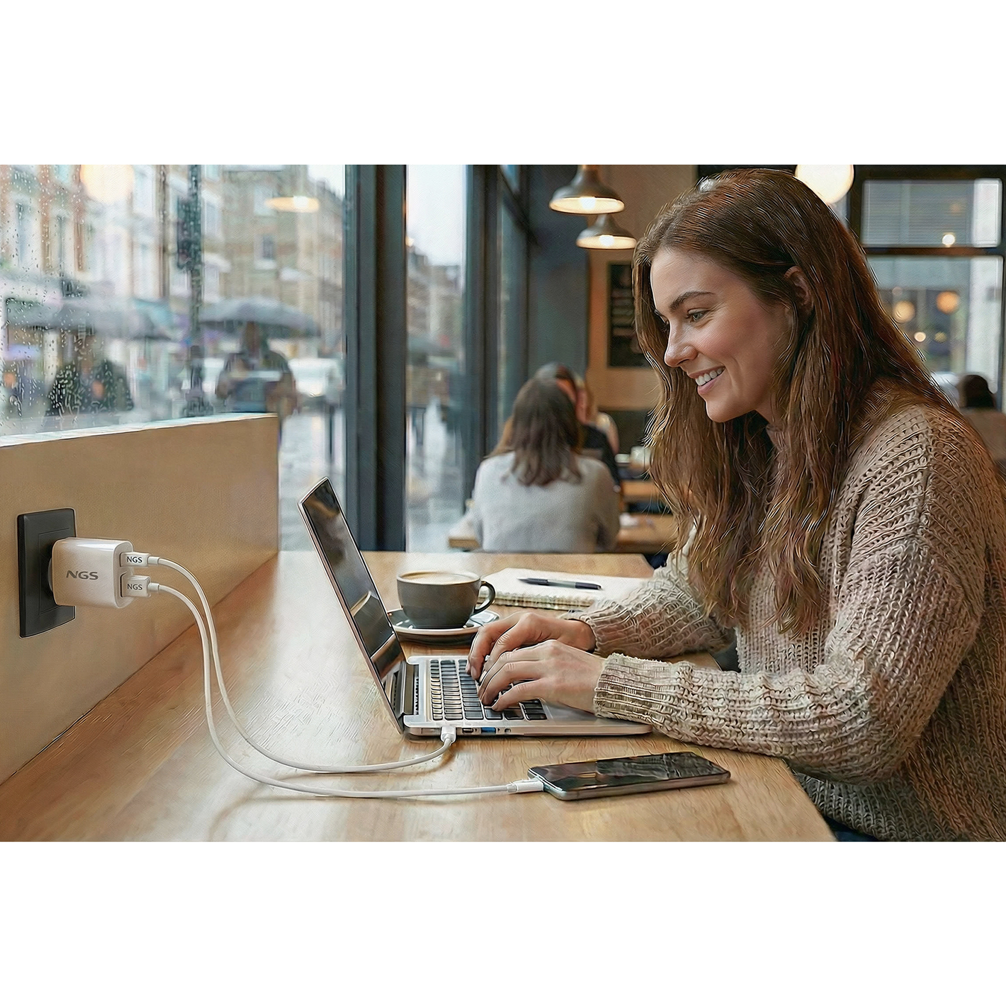 Una donna sorride mentre digita su un laptop, caricandolo e il suo telefono. Caffè e taccuino sul tavolo.