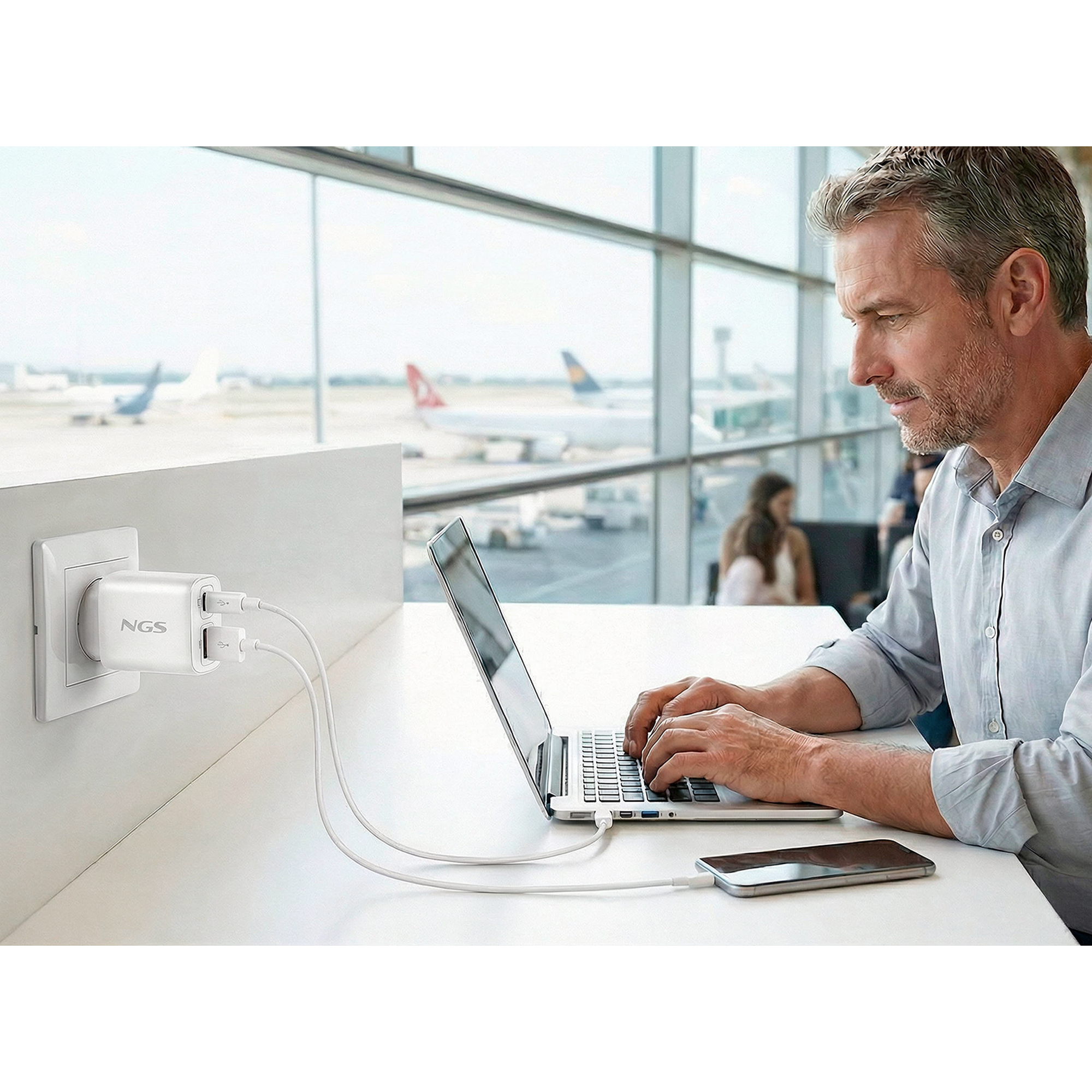 Uomo carica telefono e laptop in aeroporto con caricabatterie NGS.