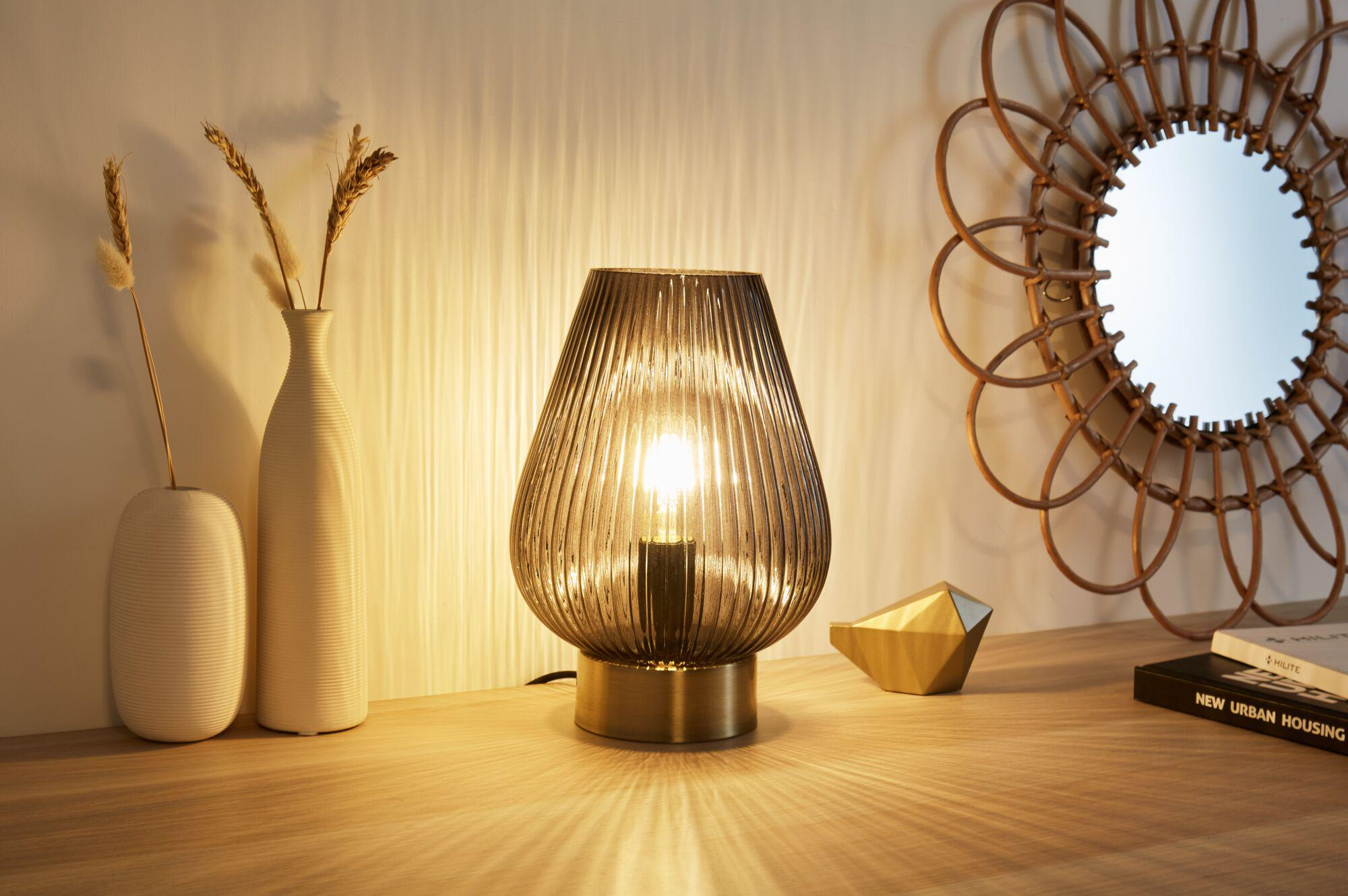 Stolik nocny z lampą, wazonami i lustrem. Ciepłe światło pada na drewniany stolik.