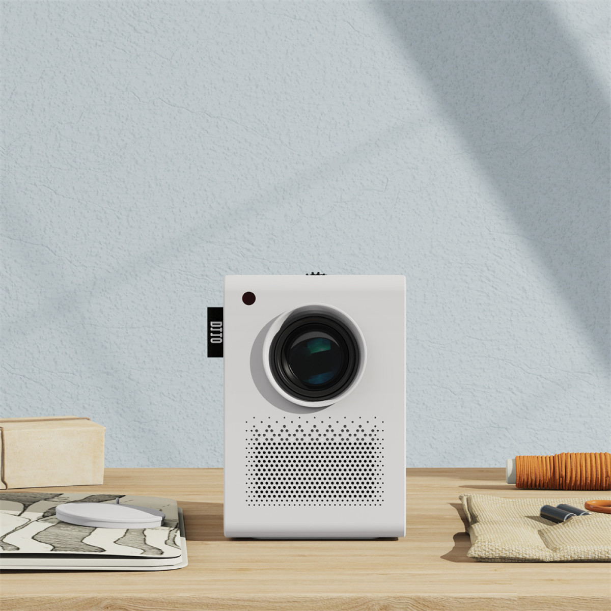 Biały projektor stoi na drewnianym stole. Po lewej pudełko, po prawej materiał. Tło jest jasnoniebieskie.