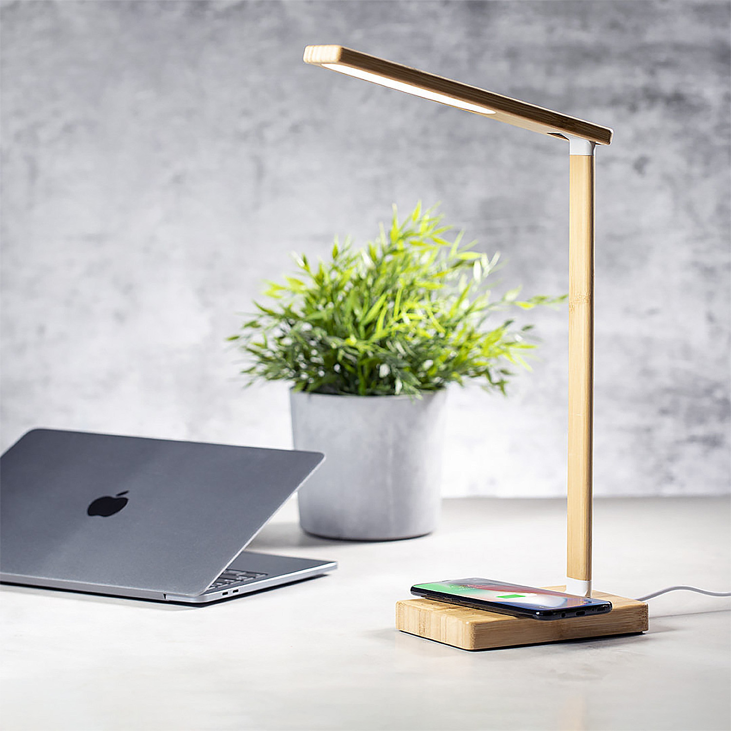 Scrivania moderna con laptop, pianta e lampada da scrivania in bambù che carica un telefono.