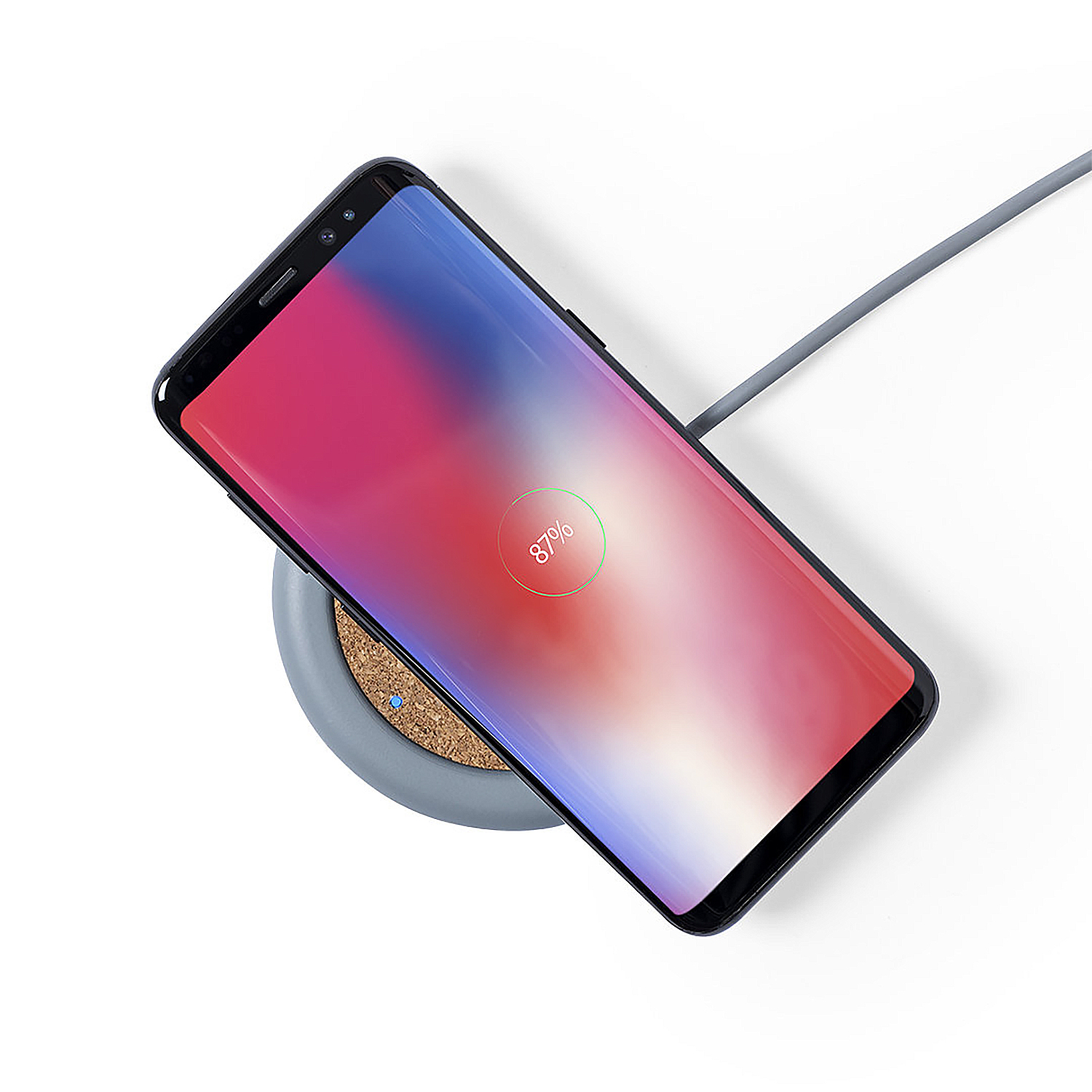 Un smartphone se carga inalámbricamente en una base de carga gris con superficie de corcho.