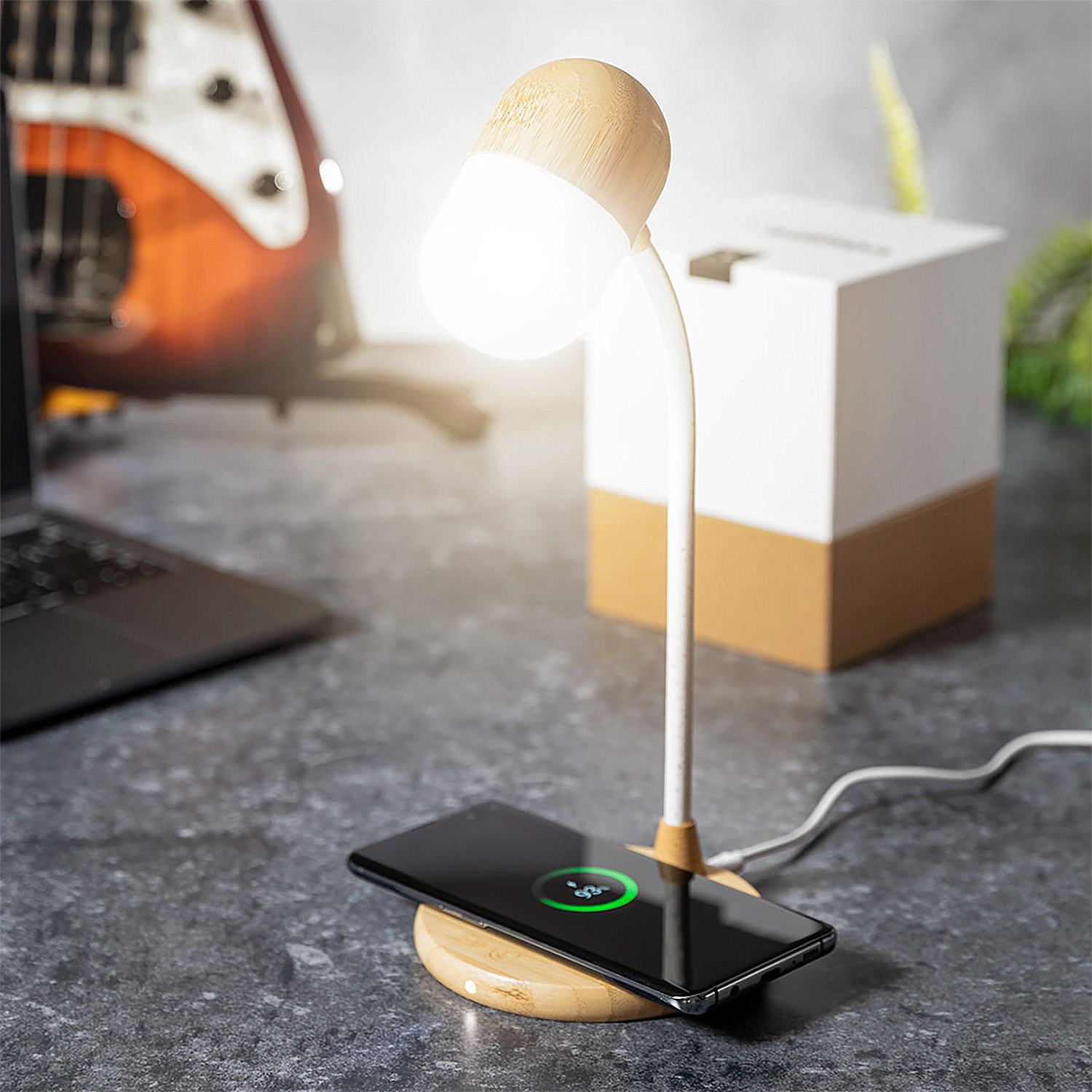 Una scrivania con lampada, telefono, laptop e chitarra. Il telefono si ricarica in modalità wireless.