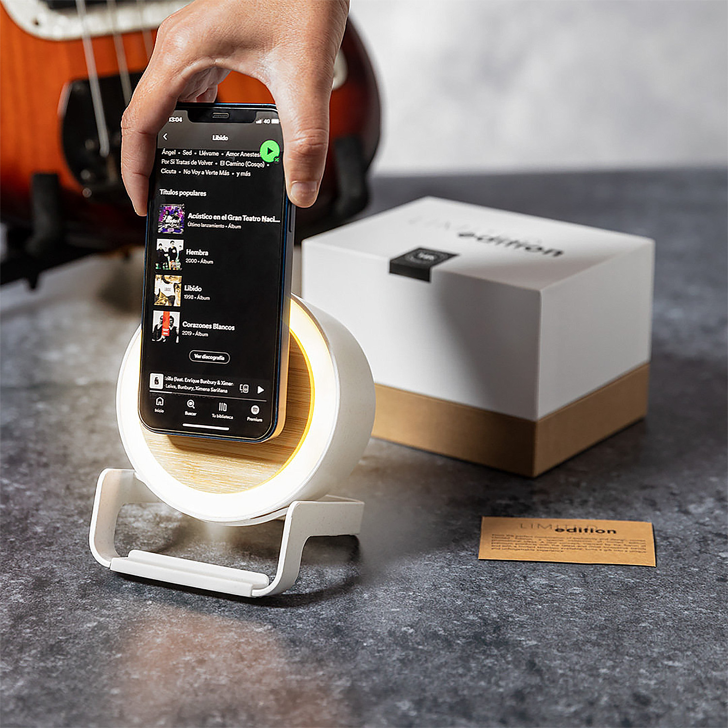Una mano sostiene un teléfono en un soporte de altavoz blanco. Una guitarra baja al fondo.