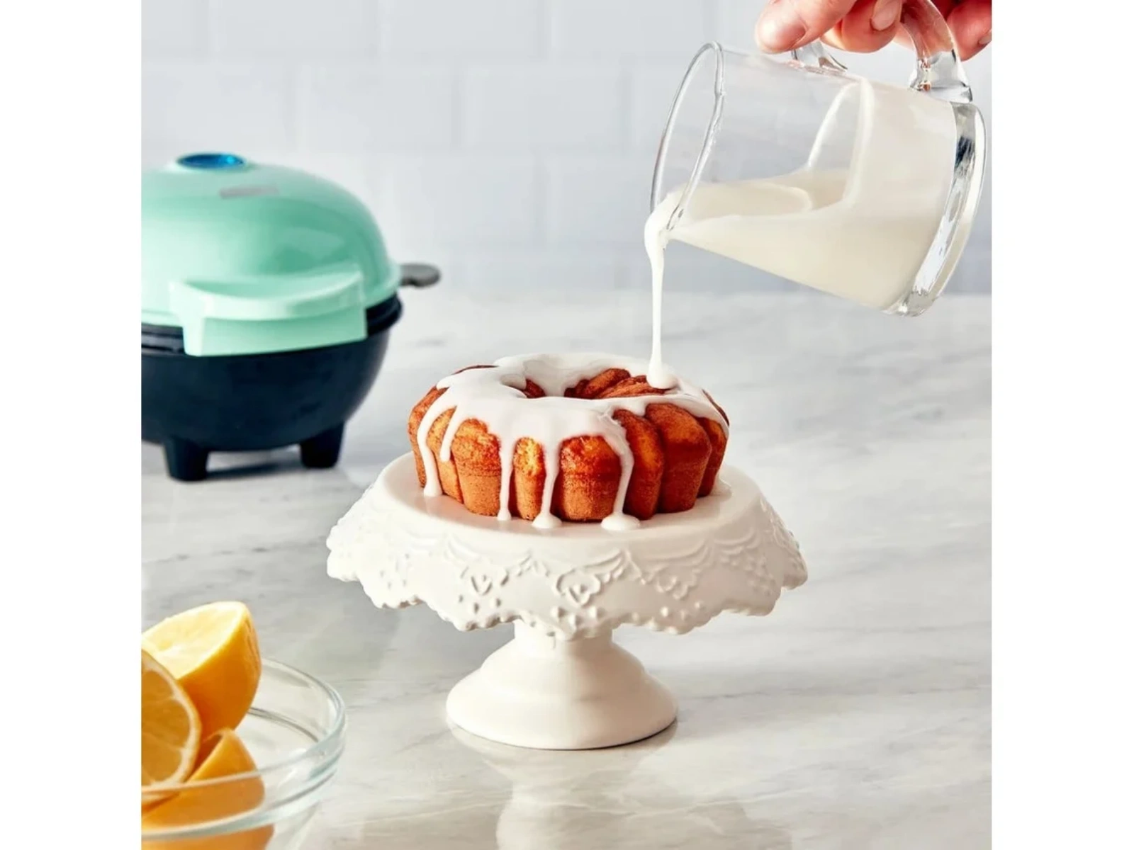 Ein kleiner Kuchen wird mit Glasur übergossen, daneben Zitronen und ein Backgerät.